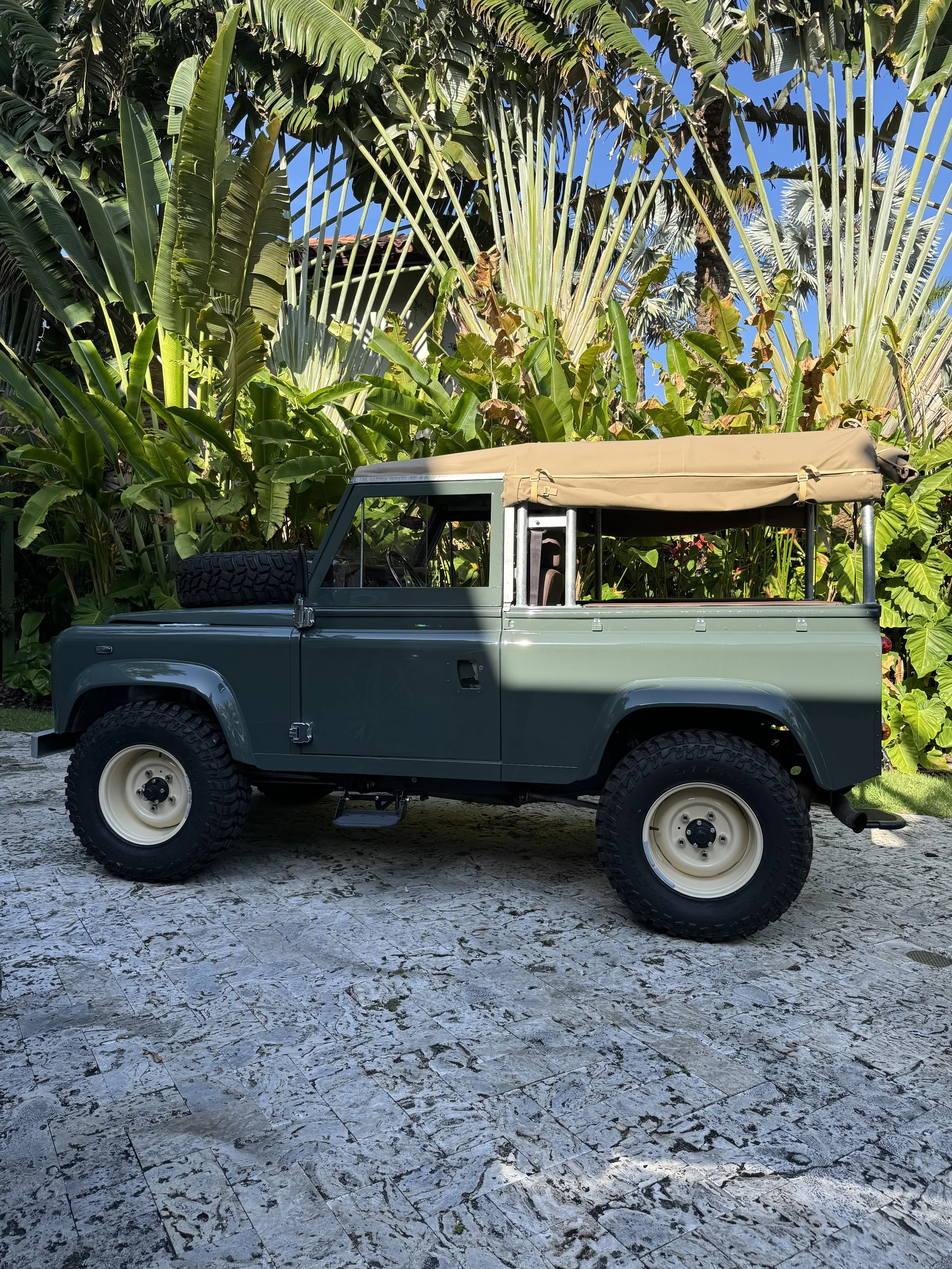 A classic green off-road vehicle with a beige soft top on a stone driveway, surrounded by lush green tropical plants.