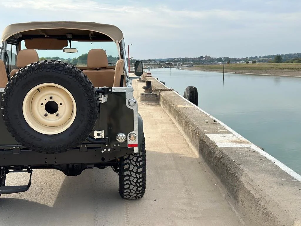 A beige off-road vehicle parked on a concrete dock beside a calm waterway with a vein, distant trees, and a cloudy sky.