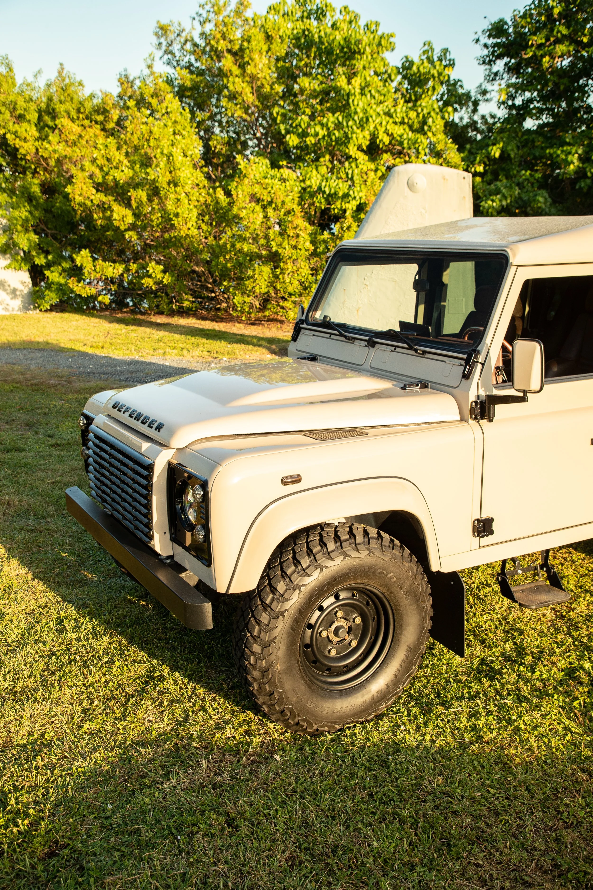 A white Land Rover Defender parked on grass with trees in the background and a clear sky.