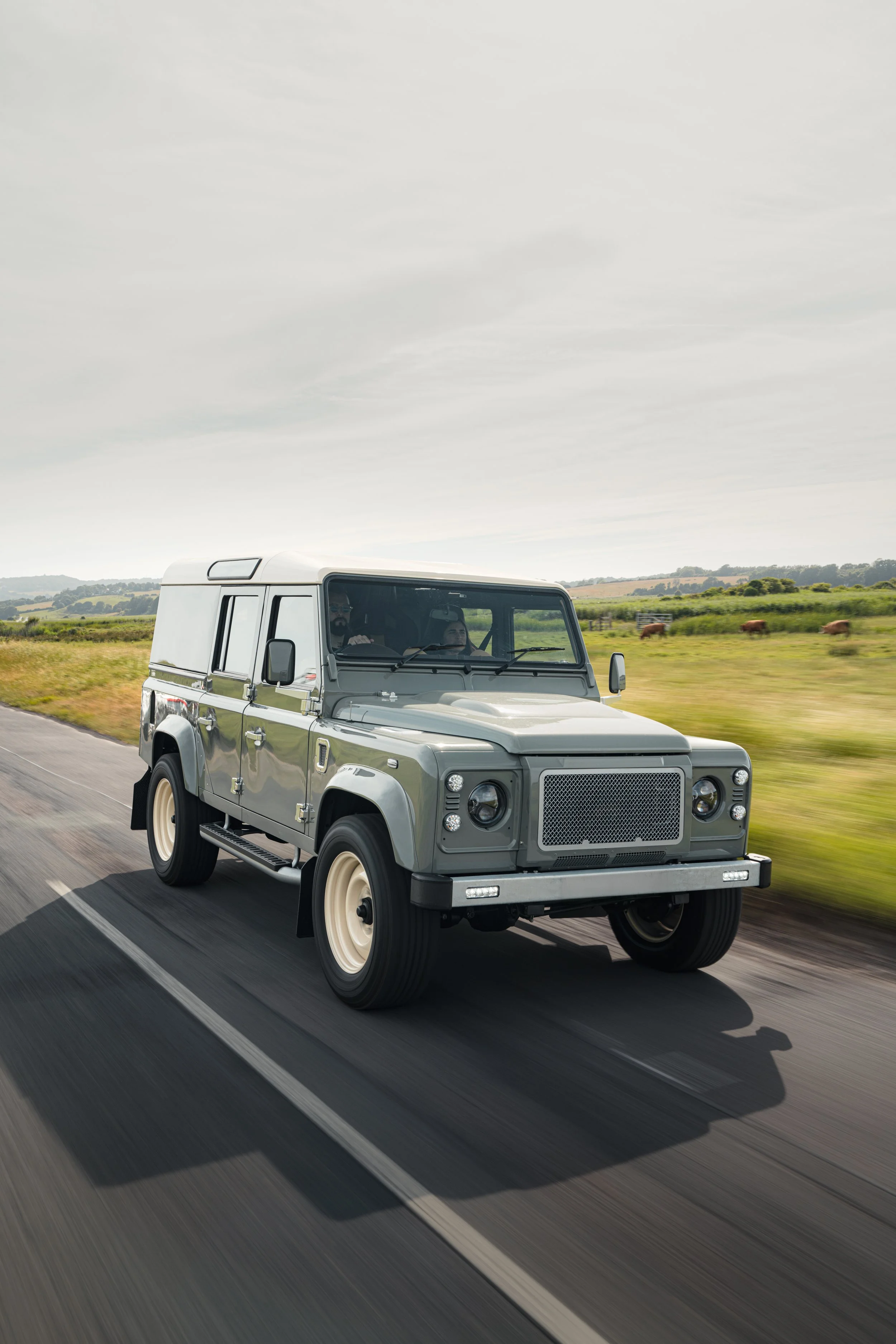 A silver Land Rover Defender driving on a rural road with fields and horses in the background.