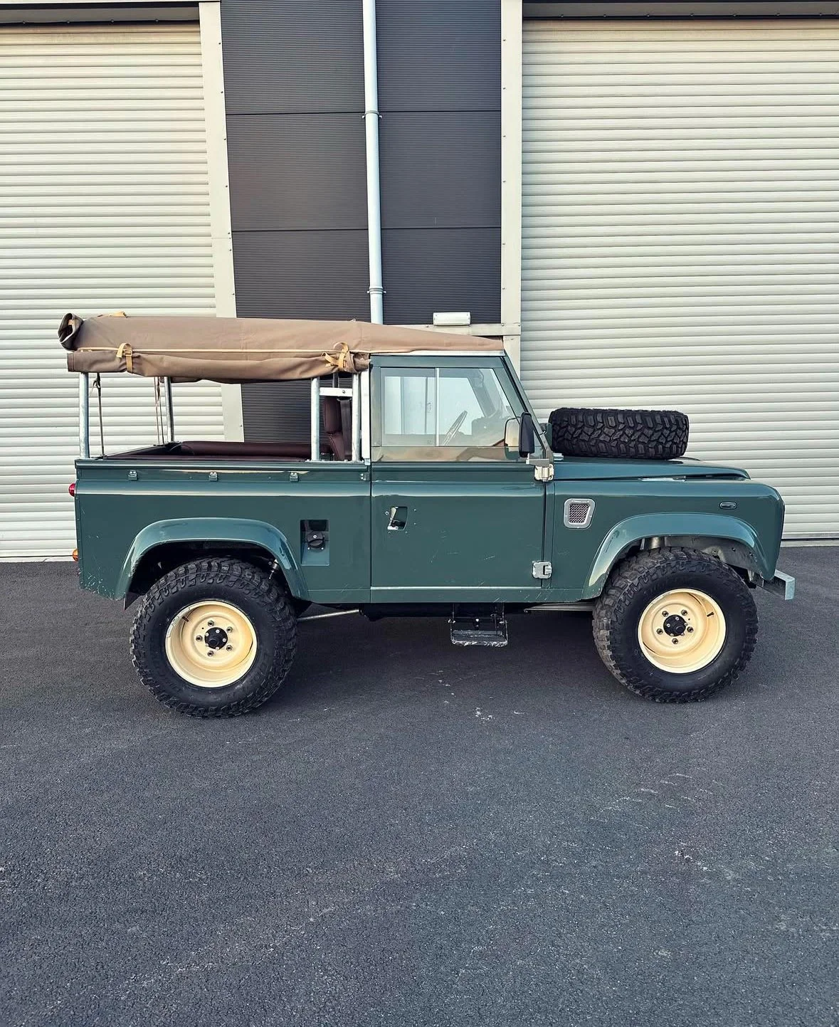 A vintage green off-road vehicle with a beige soft top and large tires parked in front of a beige and black industrial building.