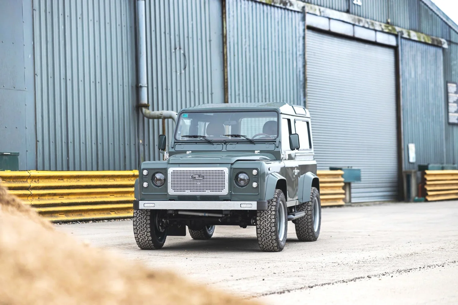 A small gray Land Rover Defender parked outside an industrial building with corrugated metal walls and yellow safety barriers.