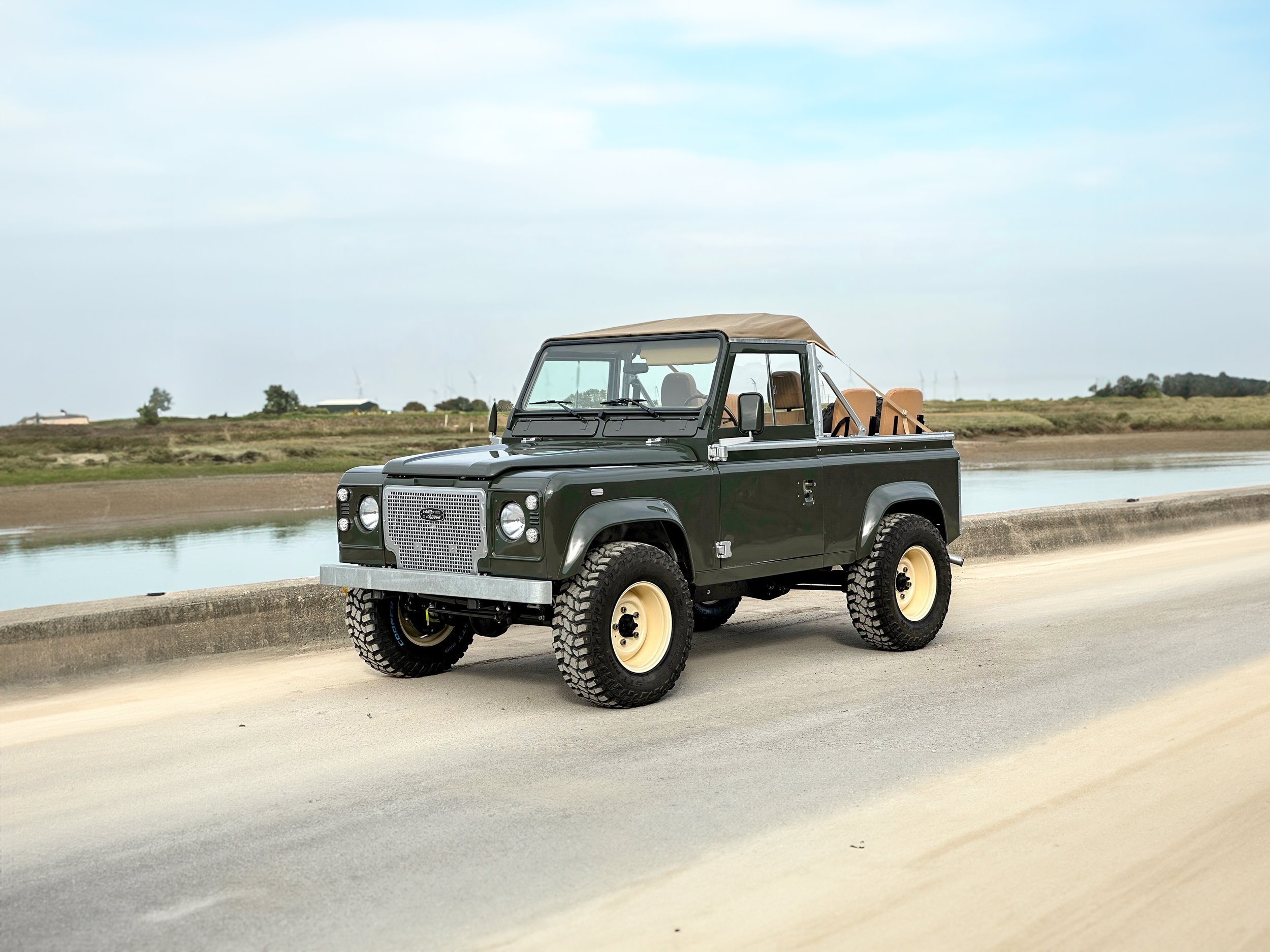 A vintage black off-road vehicle with beige seats and a tan soft top driving on a concrete road next to a body of water under a partly cloudy sky.