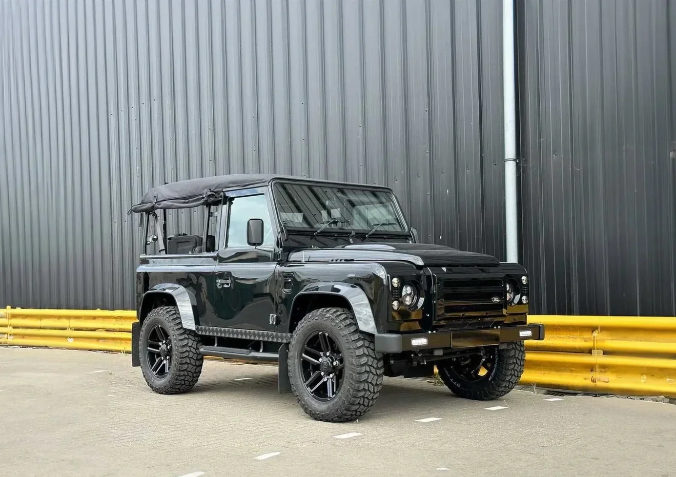 Black off-road vehicle with a soft top parked near a yellow safety barrier against a dark metal wall.