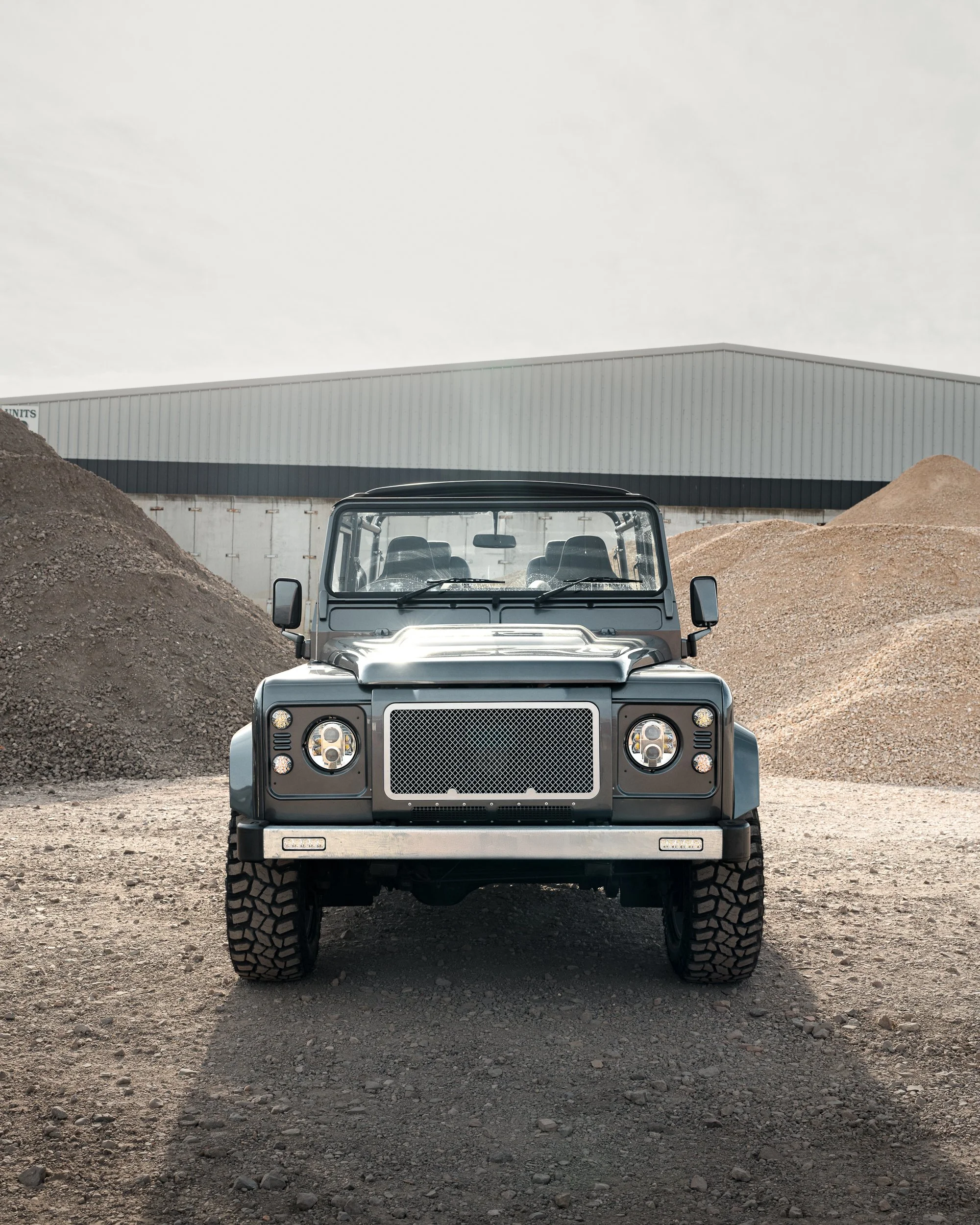 Front view of a gray off-road vehicle parked on dirt ground with piles of gravel in the background and a large industrial building behind.