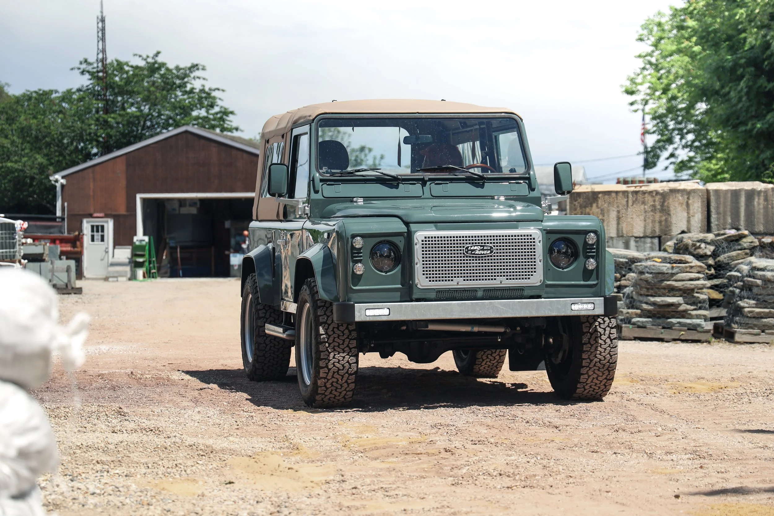 Green Land Rover Defender parked on a dirt lot with a barn in the background and stone piles to the right.