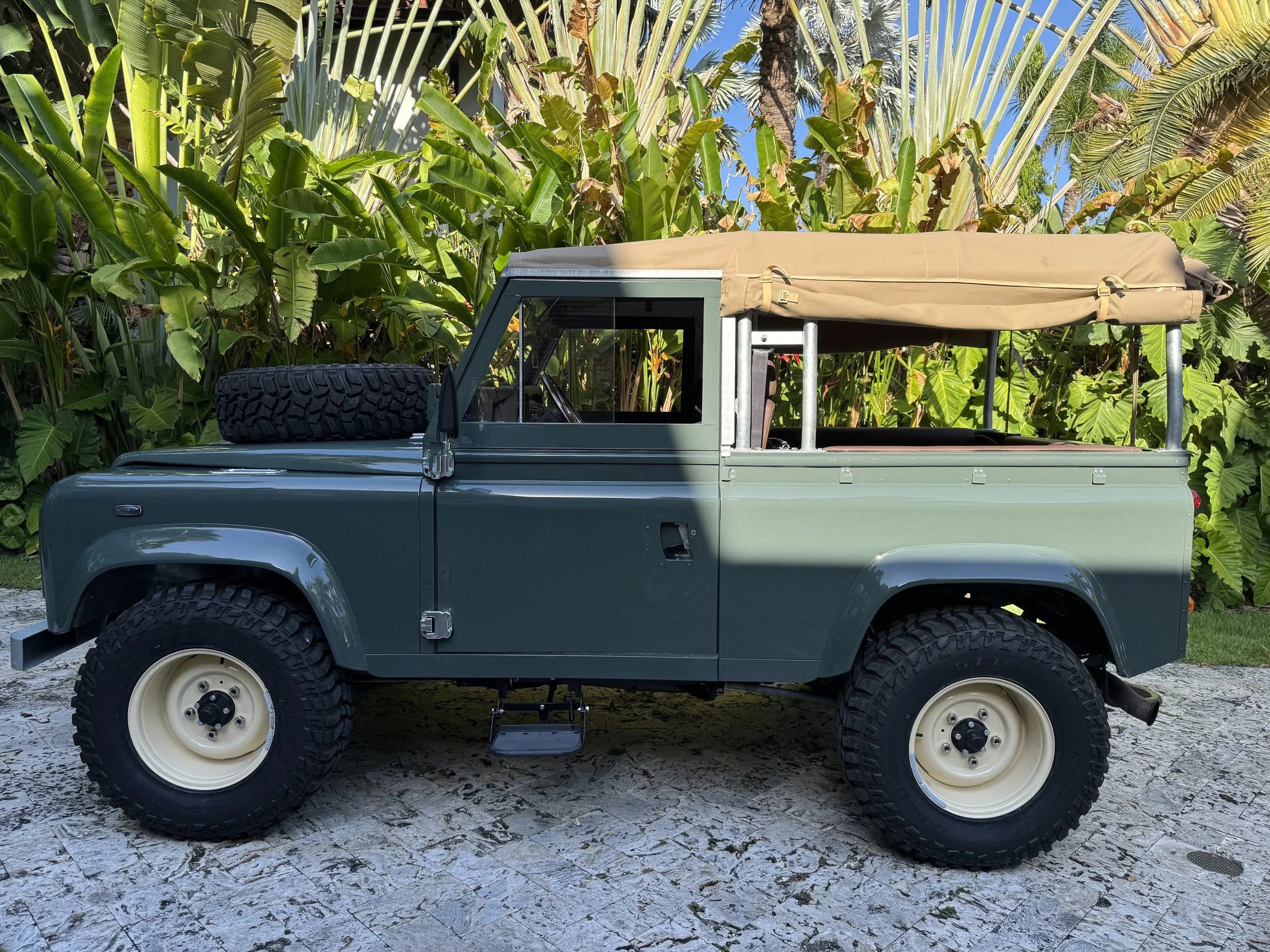 A vintage green off-road vehicle with a beige canopy, parked on a stone surface with tropical green plants in the background.