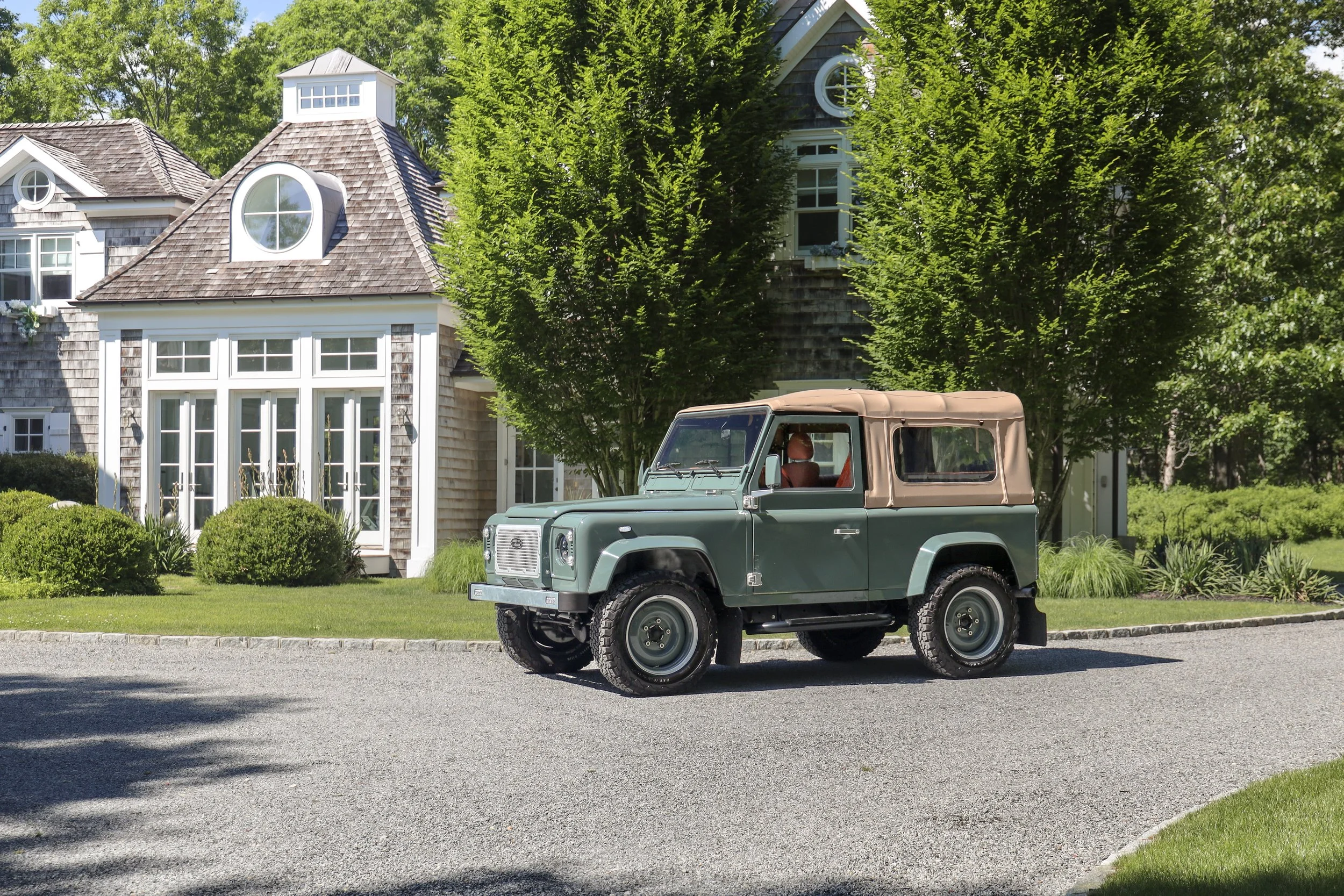 A vintage green off-road vehicle with a tan soft top parked in front of a large, elegant house surrounded by green trees and well-maintained garden.