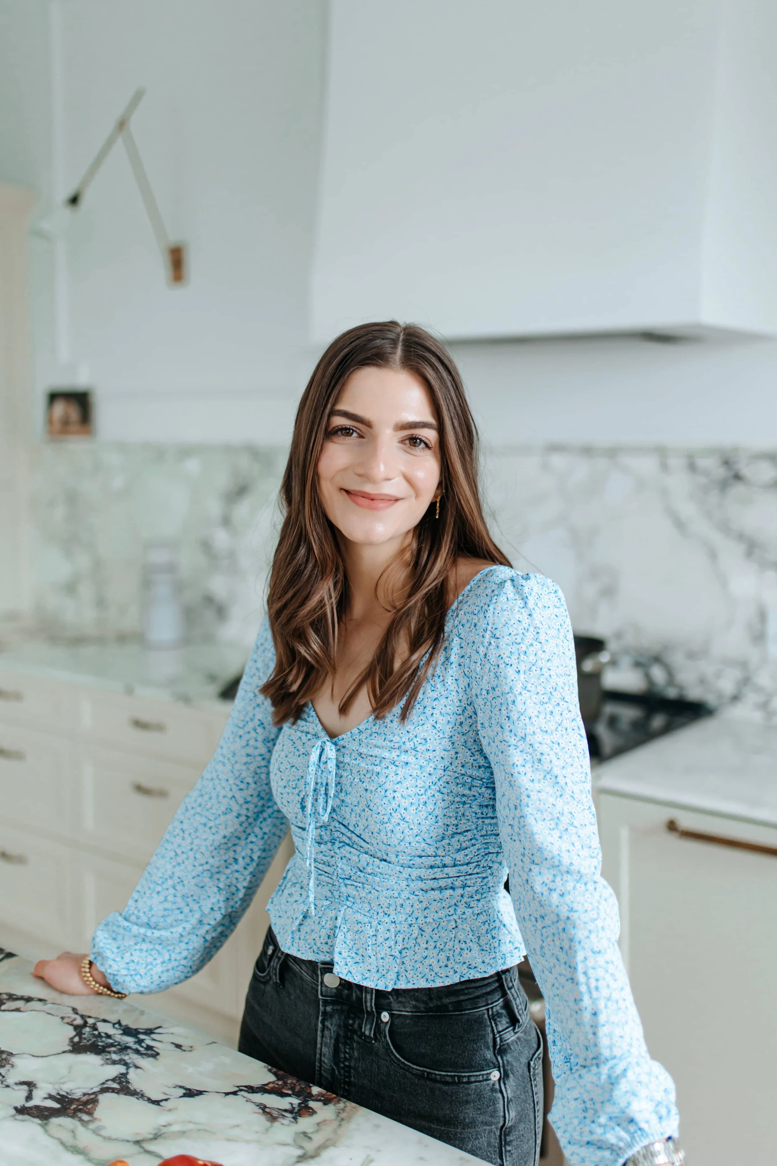 A young woman with long brown hair, wearing a light blue floral blouse and black jeans, smiling and leaning on a marble kitchen countertop.