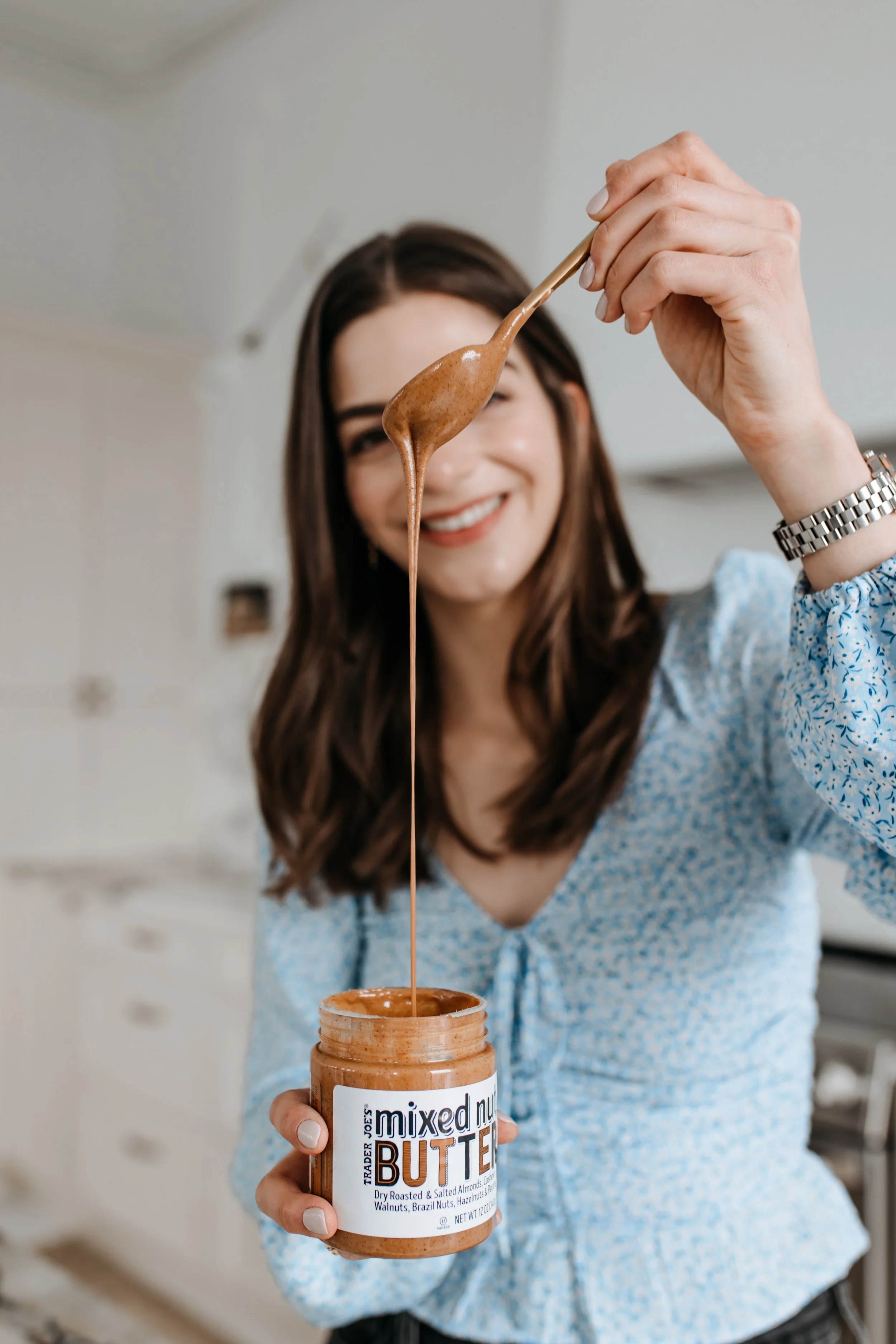 A woman in a light blue blouse smiling and holding a jar of mixed nut butter, with some nut butter on a spoon she is lifting