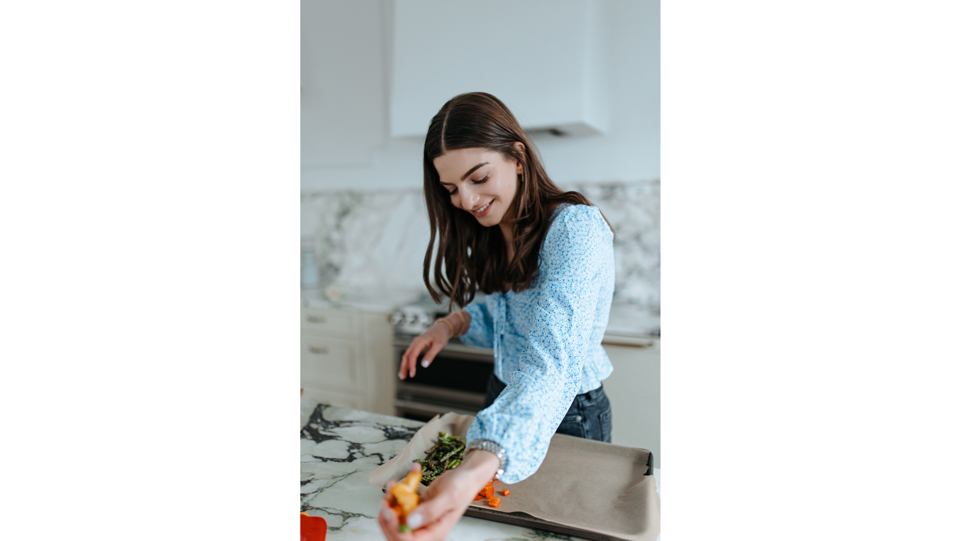 A woman in a blue patterned blouse preparing vegetables on a marble kitchen countertop, smiling while placing chopped vegetables onto a baking sheet lined with parchment paper.