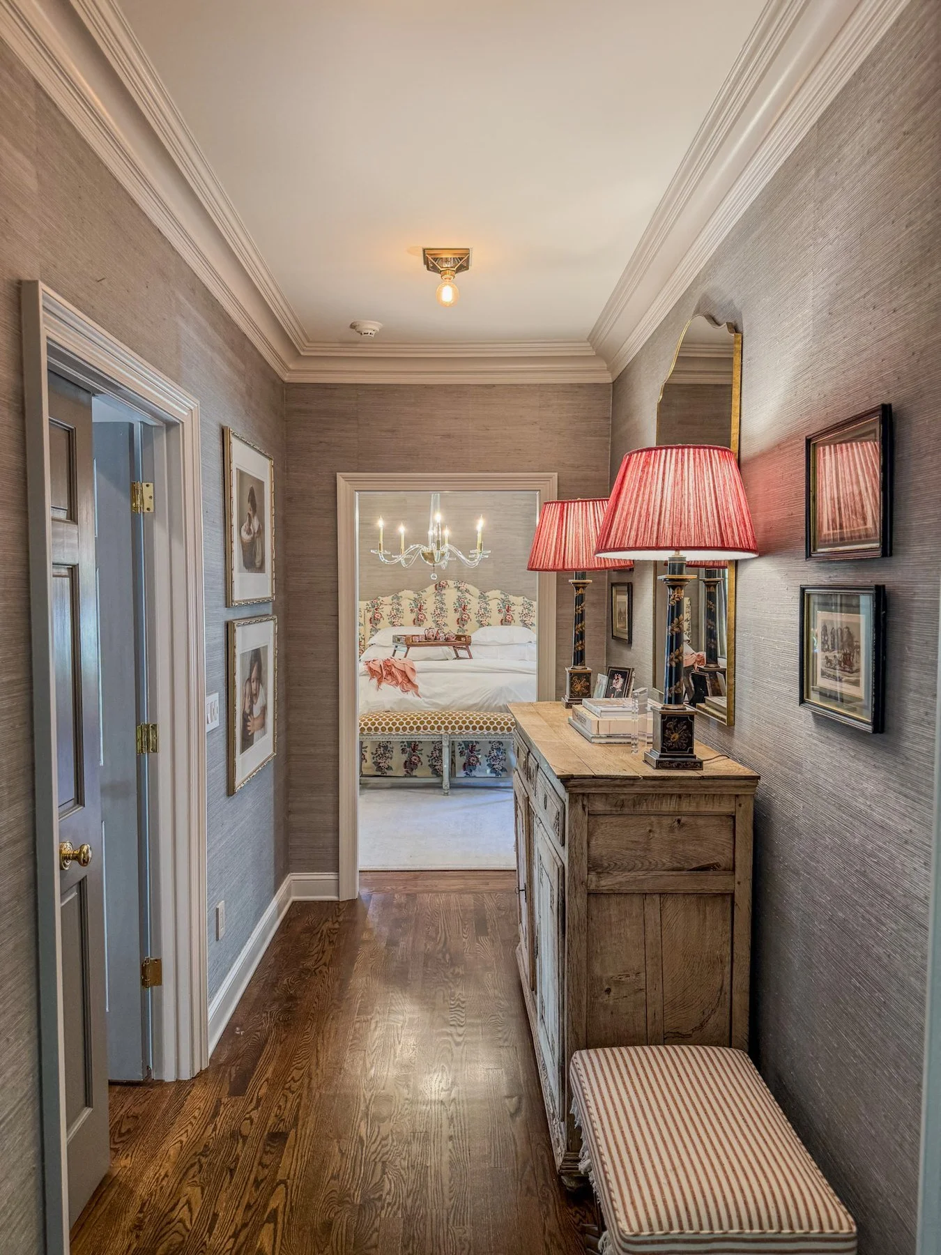 Grasscloth-walled hallway with gallery wall and antique console leading into a floral-canopied bedroom