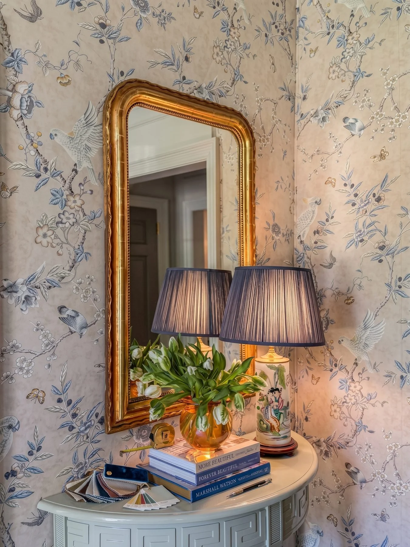 Chinoiserie floral wallpaper with a gold-framed mirror, navy pleated lamp, and white tulips on a console.