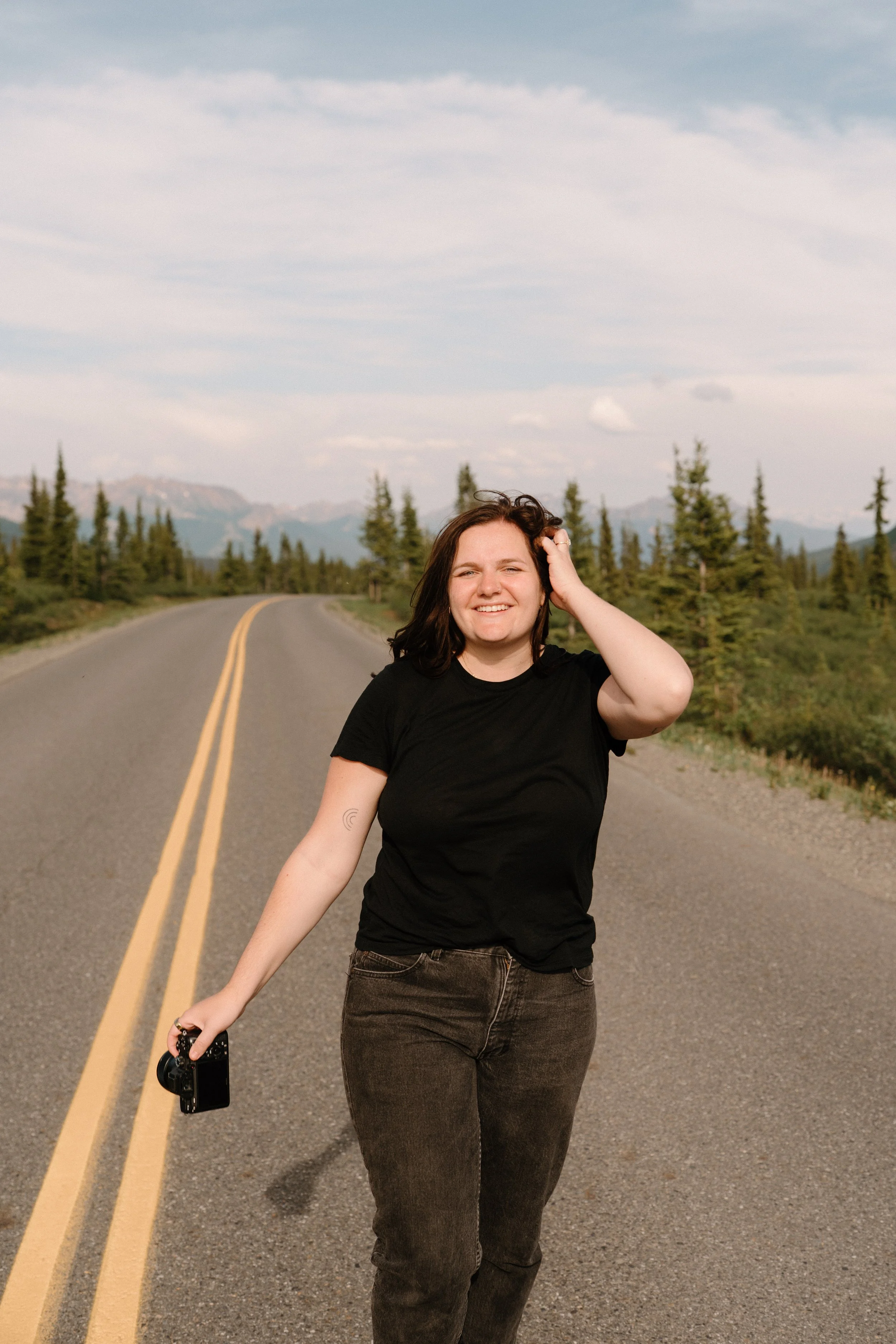 A woman walking on the road with greenery and mountains in the background, smiling and wearing a black t-shirt, holding a camera in her right hand.