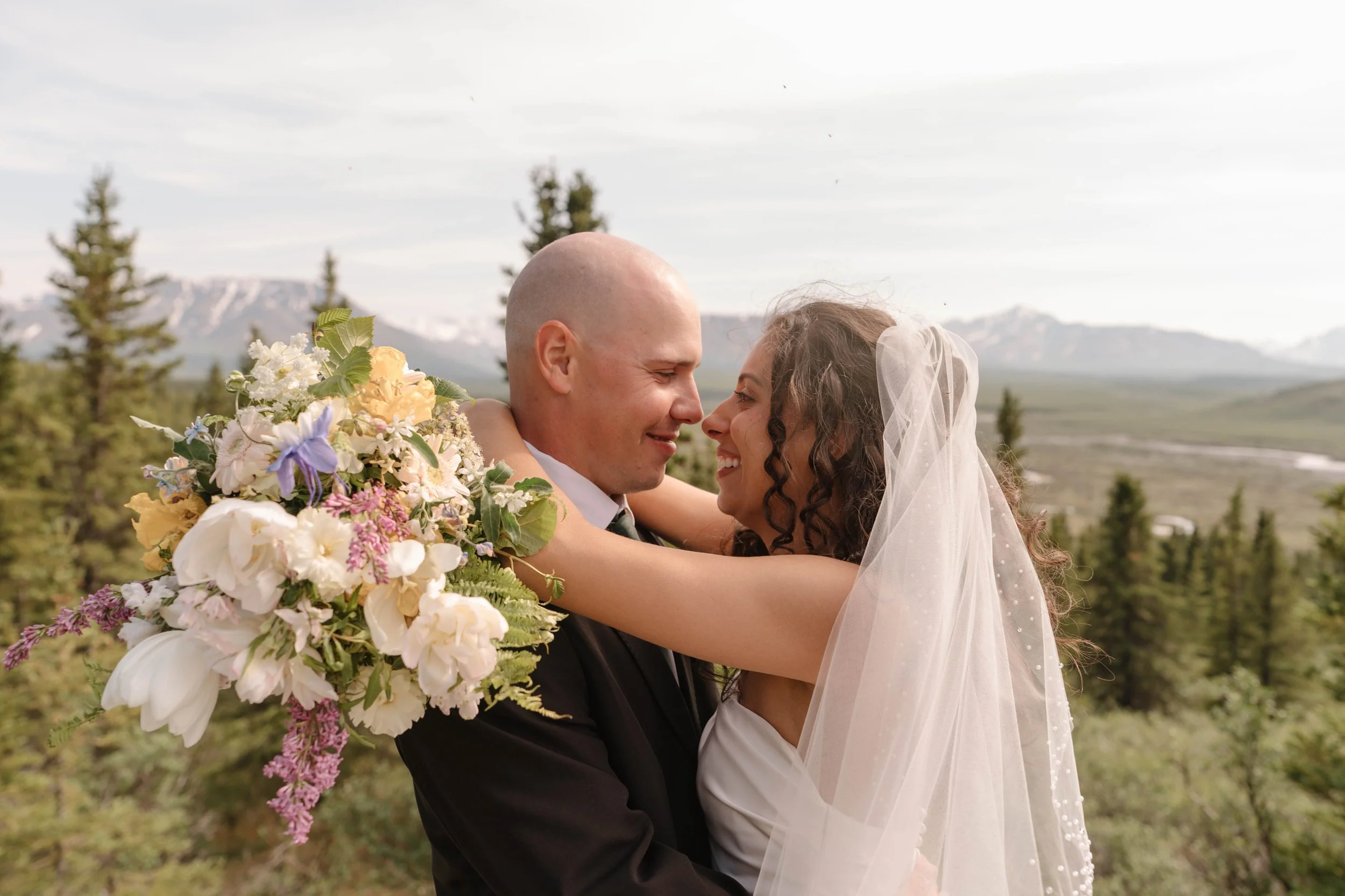 A bride and groom embrace outdoors with mountains and trees in the background. The bride is wearing a white wedding dress and veil, and is holding a large bouquet of flowers. The groom is in a dark suit, posing in Denali National Park in Alaska