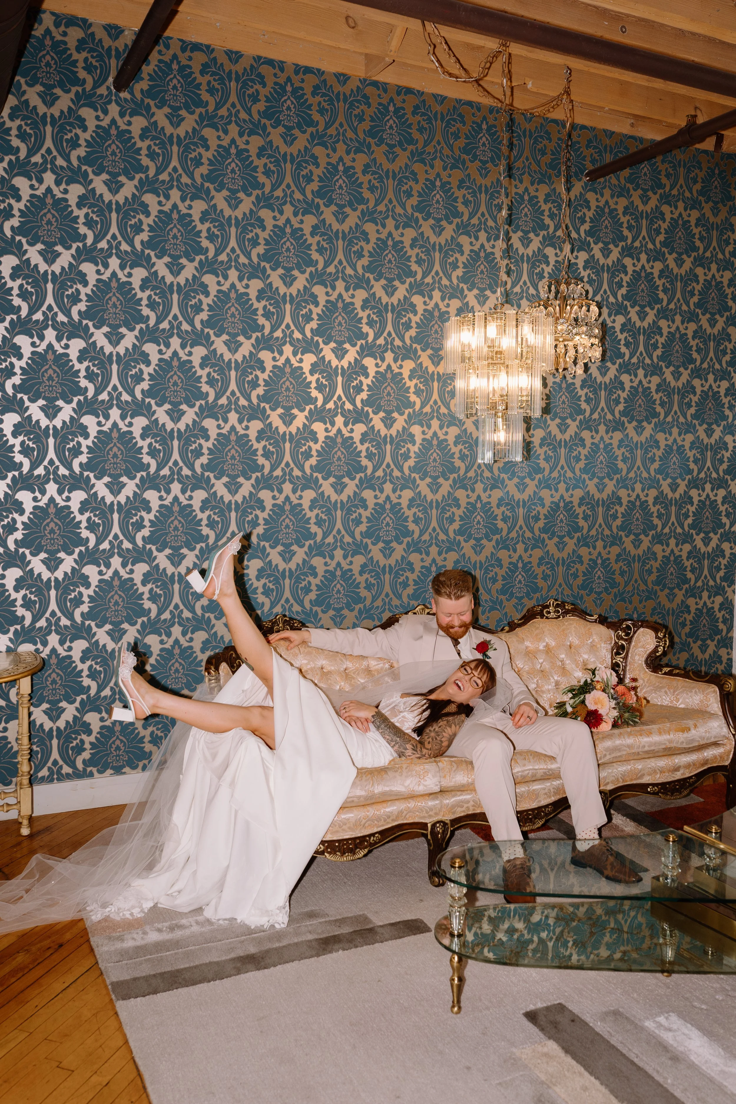 A newlywed couple is sitting on a vintage sofa, with the groom laughing and the bride smiling and lifting her leg. The bride is wearing a white wedding dress and glasses, and the groom is in a beige suit with a red boutonniere. A bouquet of flowers is resting on the sofa, and a chandelier hangs above them against a blue damask patterned wall.