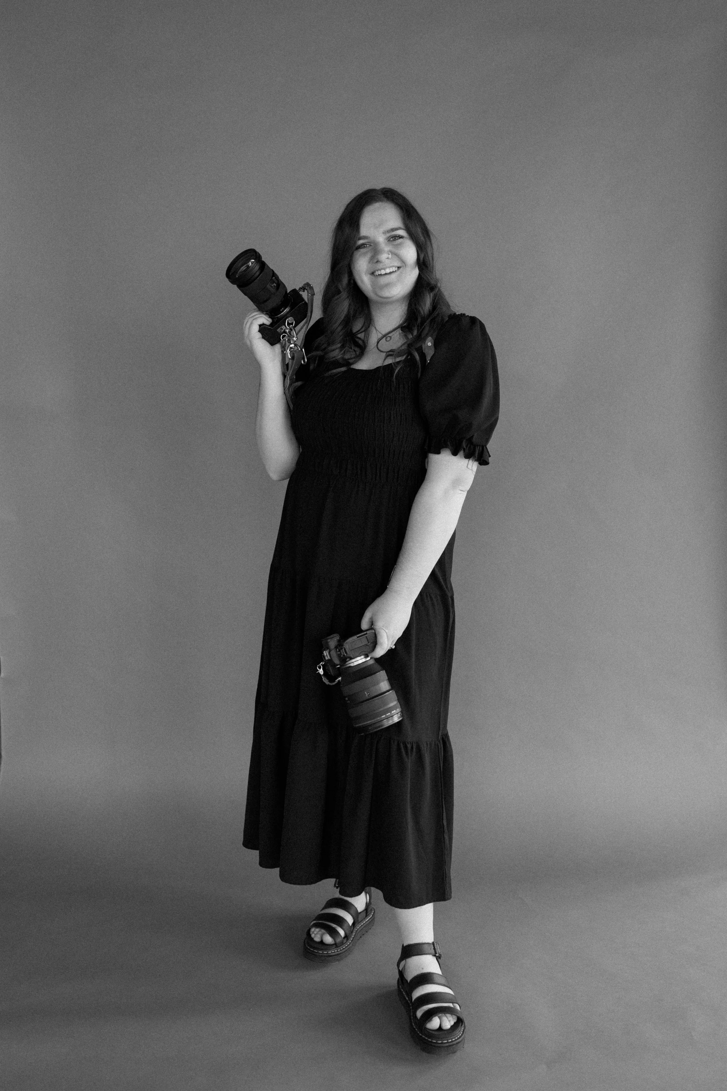 A woman with long dark hair is smiling while holding two cameras, one in each hand, standing against a plain background.