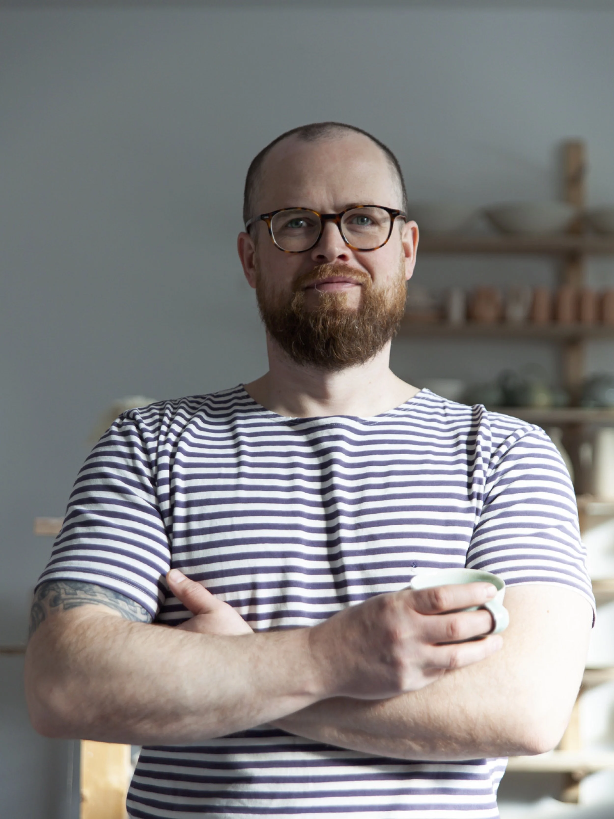 Ein Mann mit Brille und Vollbart, trägt ein gestreiftes T-Shirt, hält eine Tasse.