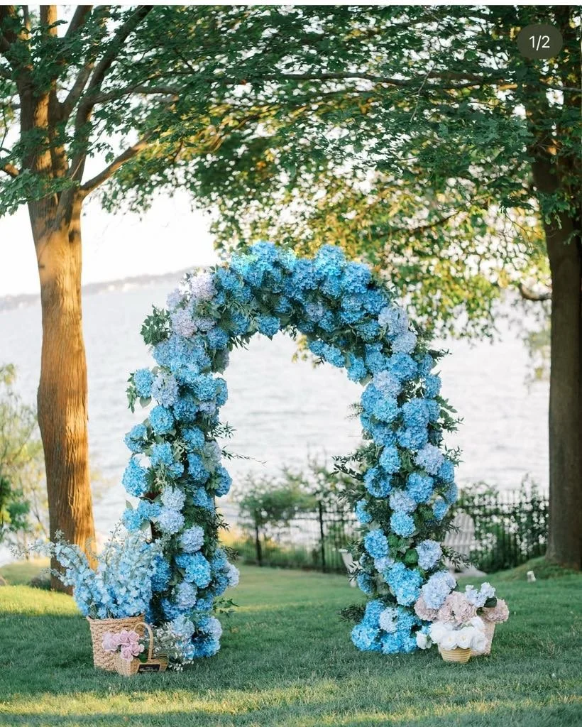 Blue and white flower arch outdoors near water, with baskets of flowers at the base and trees in the background.