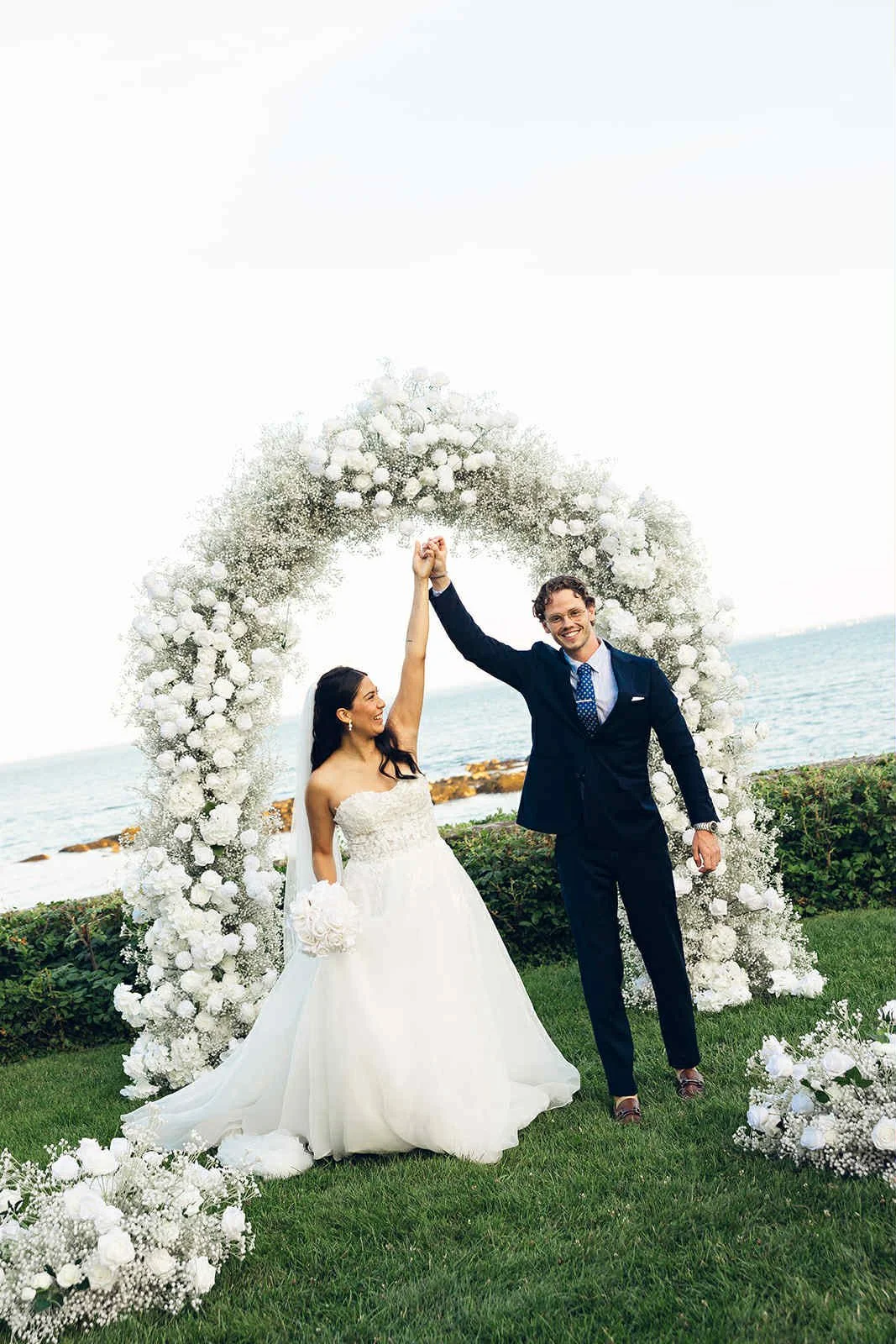 A newlywed couple celebrating outdoors by the ocean under a floral arch, with the bride in a white wedding gown holding a bouquet and the groom in a dark suit, holding hands raised in victory.