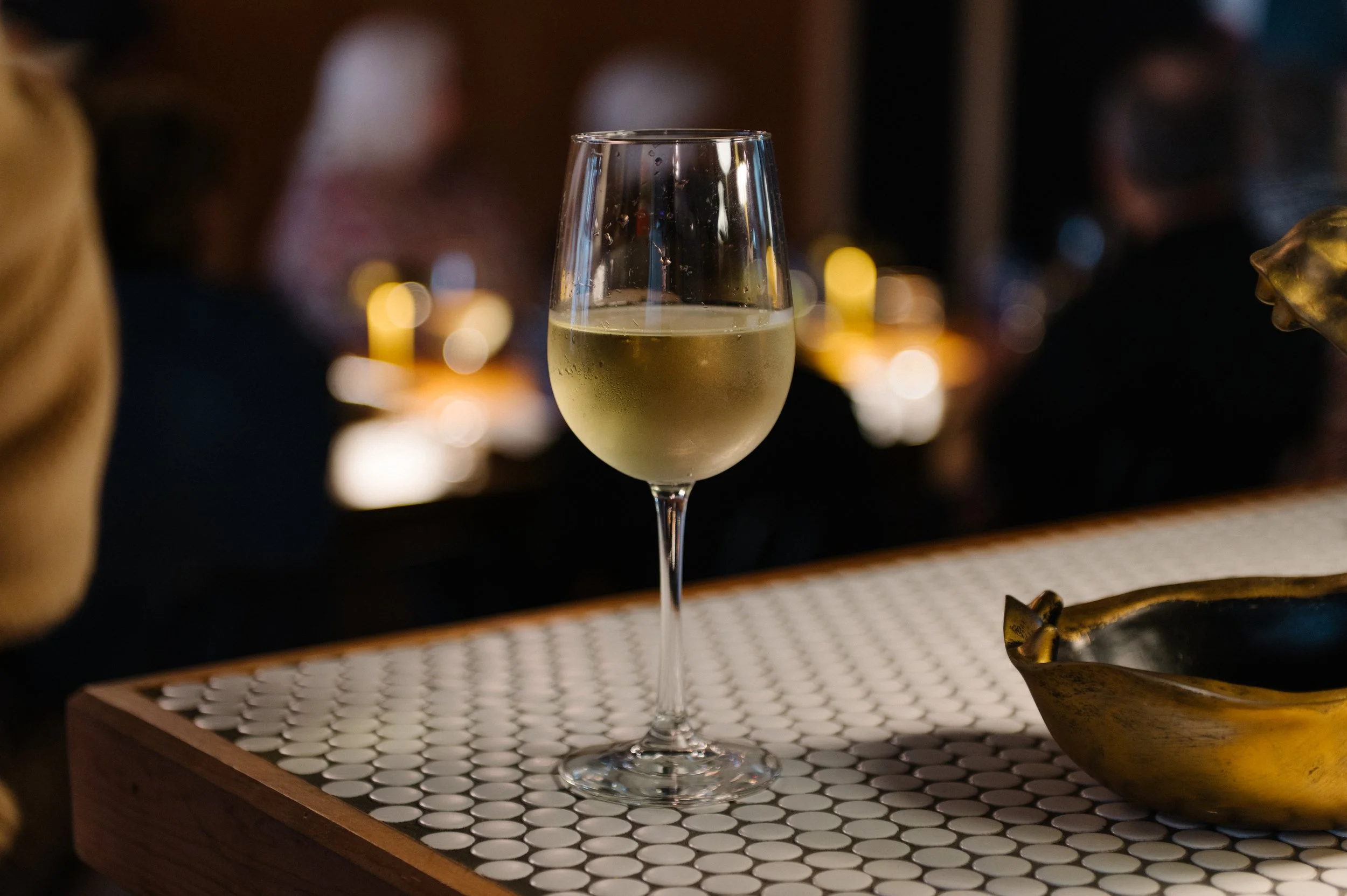 A glass of white wine on a white-tiled table with a brass bowl nearby, in a dimly lit setting with a blurred background of people and warm lights.