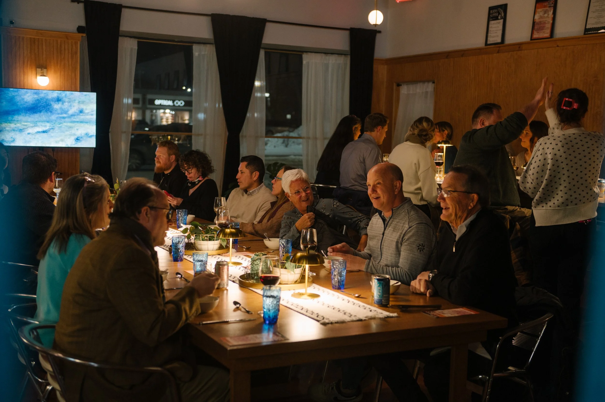 A group of people sitting at a long dining table in a warmly lit restaurant, engaged in conversation and laughter, with some standing at the bar in the background. Des Moines Team Building.