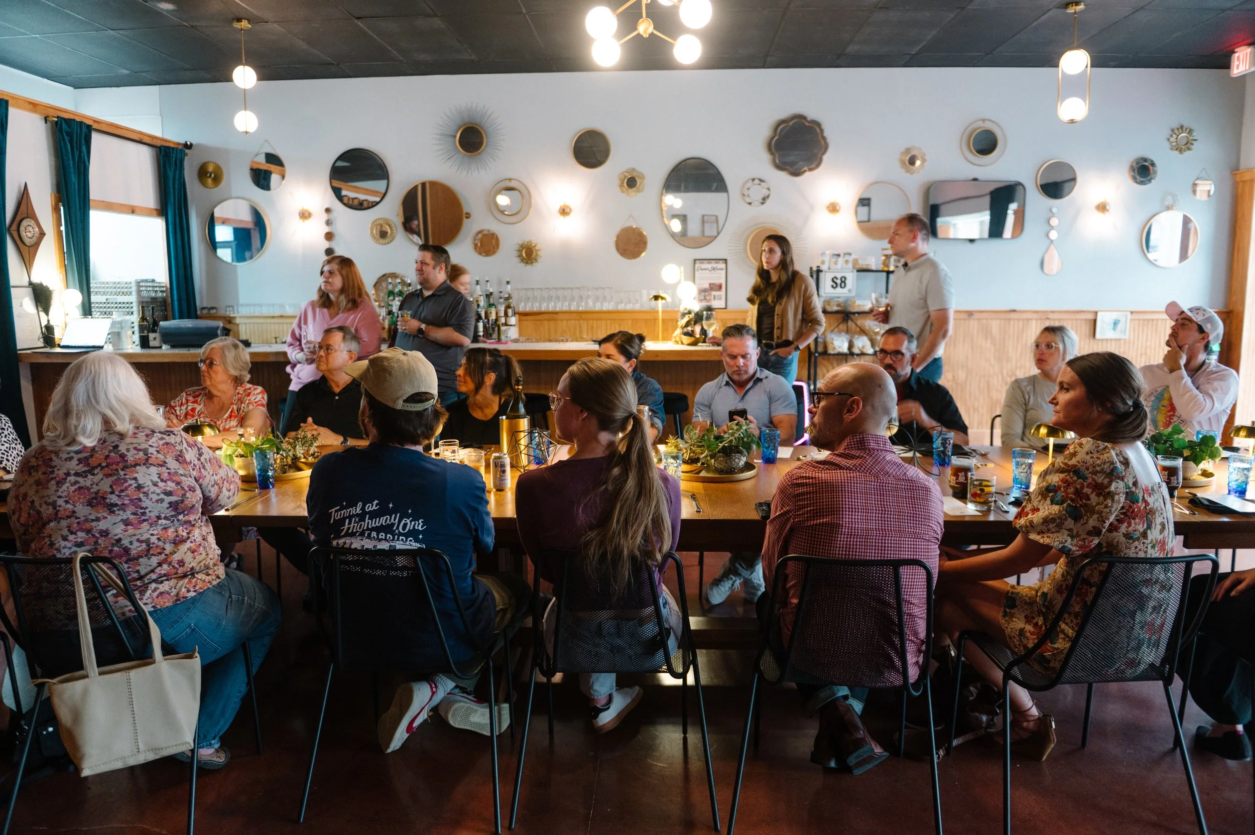 A group of people seated around a long dining table in a restaurant or cafe, with some standing by a bar in the background. The space is decorated with various round mirrors and decorative wall pieces, with warm lighting and a cozy atmosphere.