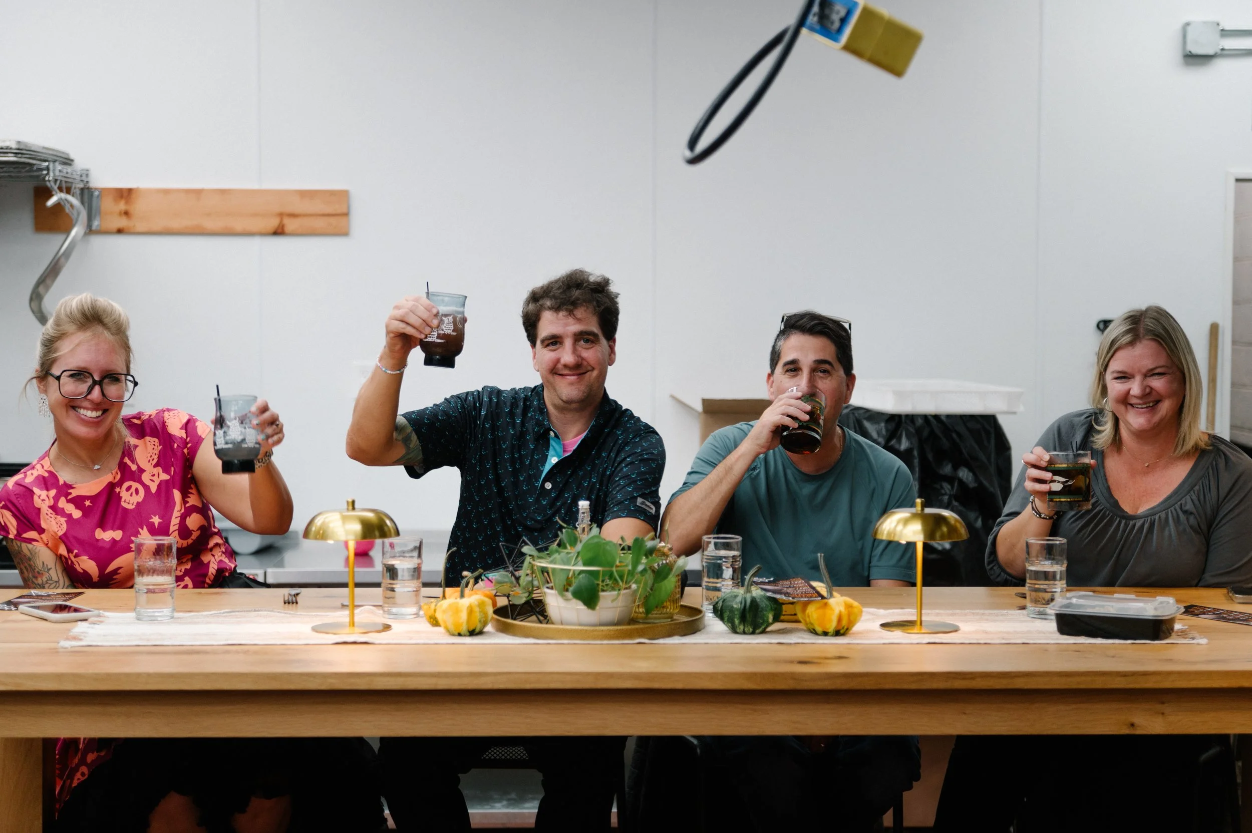 Four people sitting at a dining table, smiling, and raising drinks for a toast indoors, with a centerpiece and small decorative pumpkins on the table.
