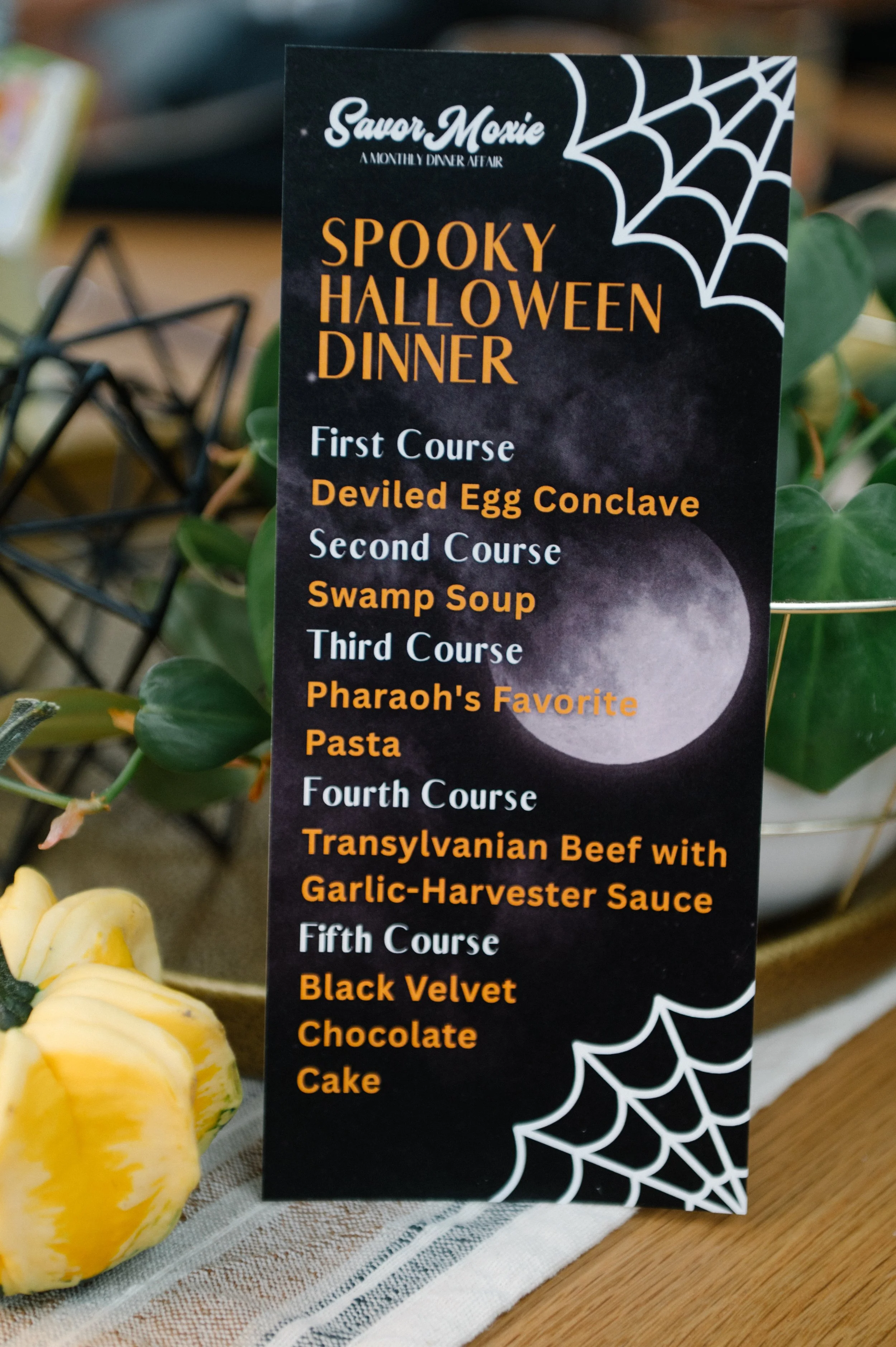 A Halloween-themed menu featuring a spooky moon and spiderweb design, listing a five-course Halloween dinner including deviled egg conclave, swamp soup, Pharaoh's favorite pasta, transylvanian beef, and a black velvet chocolate cake.