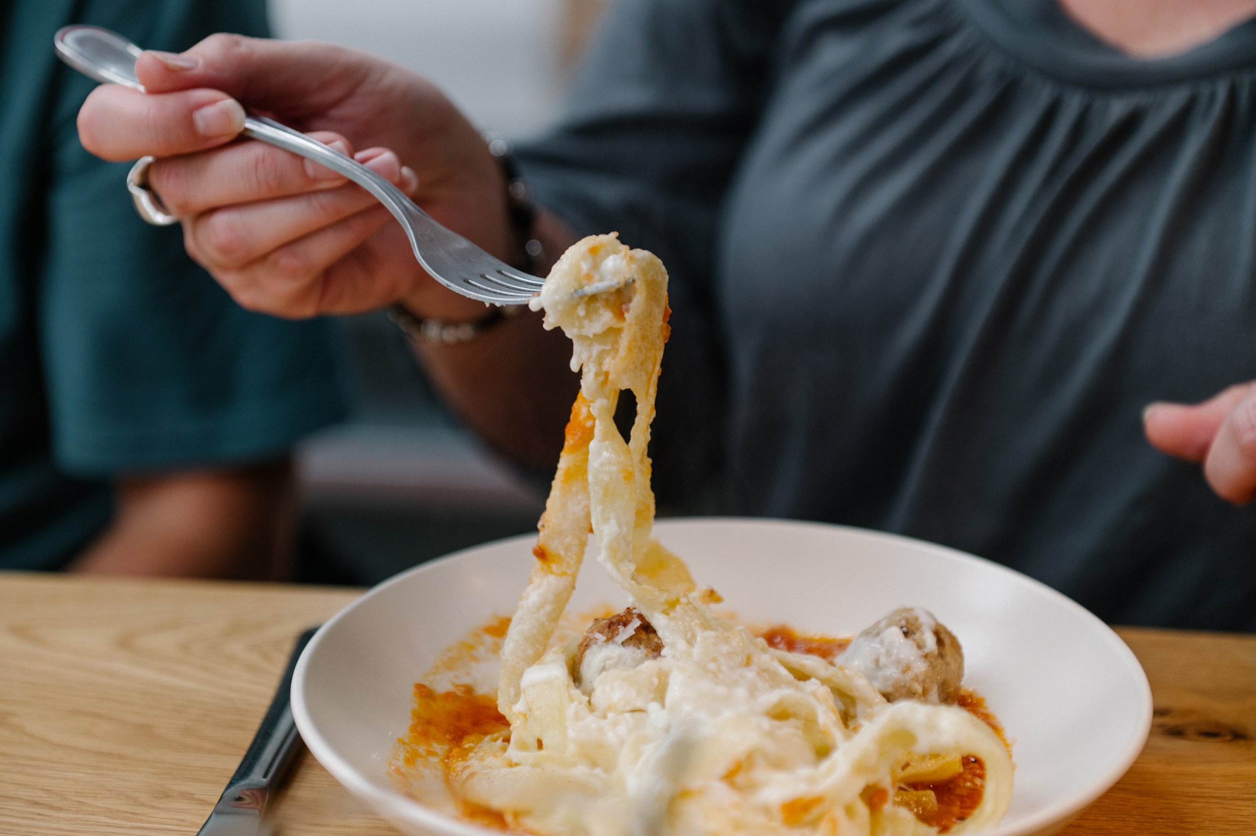 Person holding fork with a bite of cheesy lasagna, served on a white plate on a wooden table.