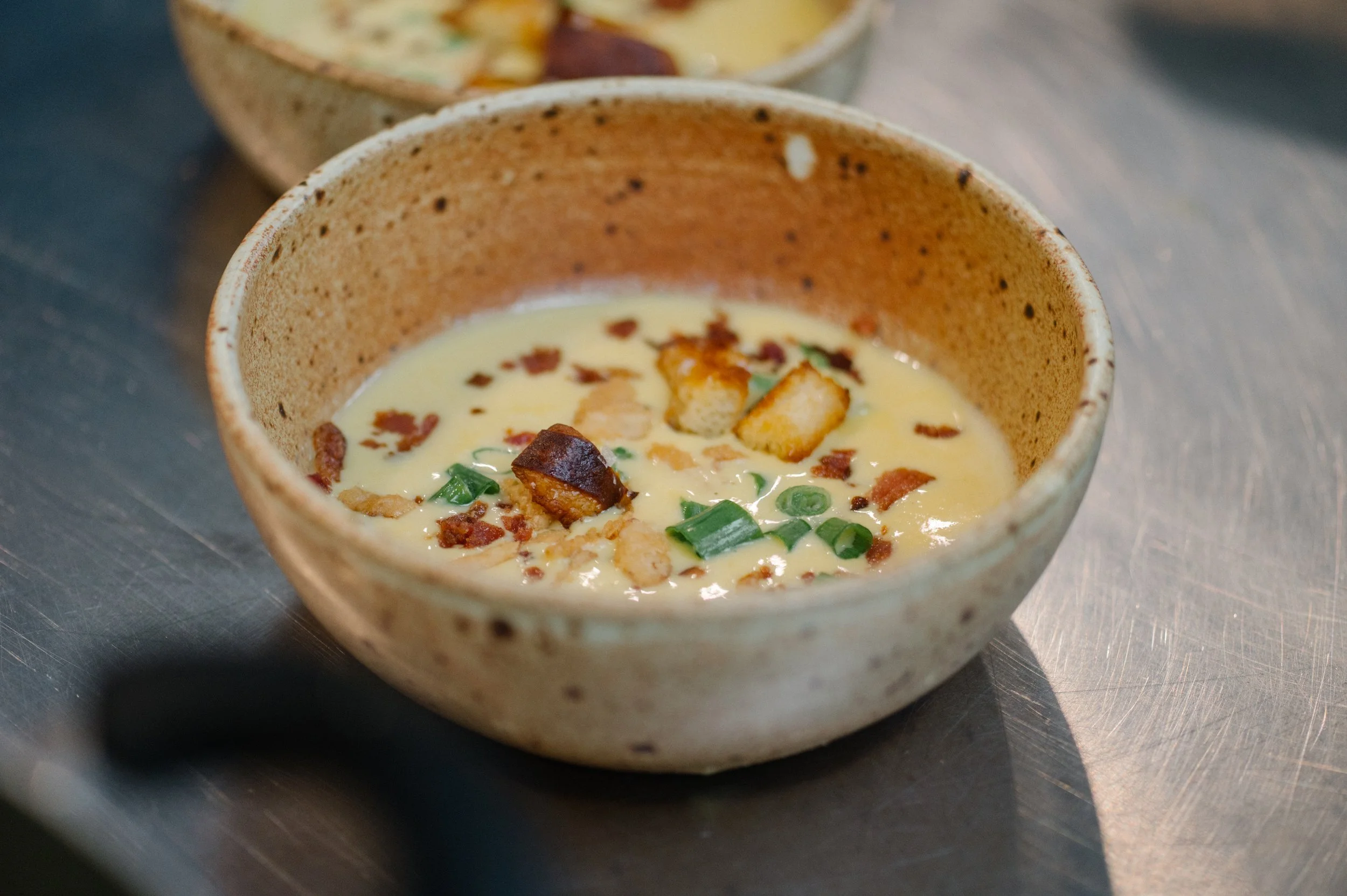Creamy soup in a rustic ceramic bowl topped with croutons, chopped green onions, and crispy bacon bits on a metal surface.