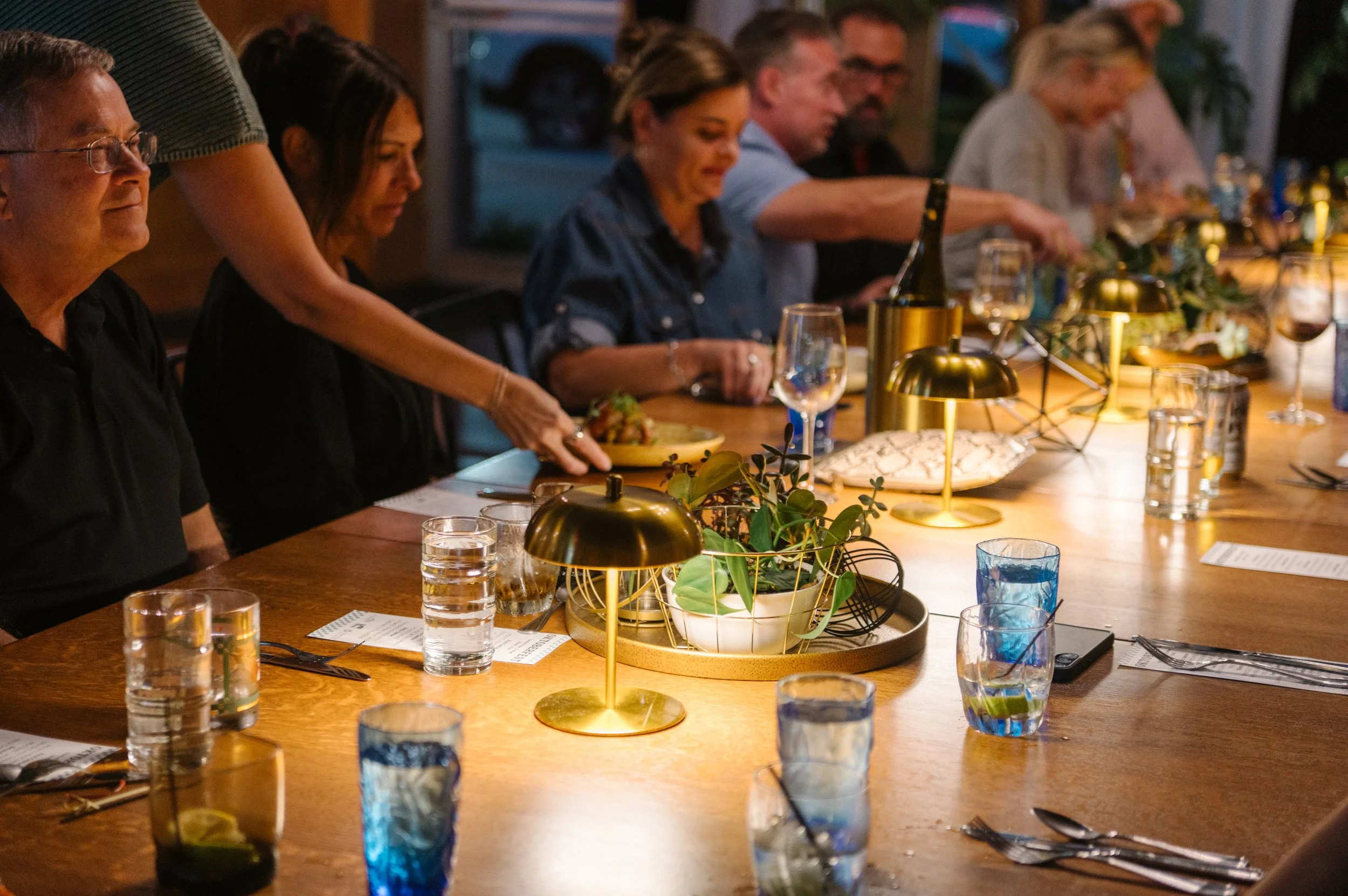 People seated at a dinner table with glasses of water, wine, and a bottle of champagne, decorated with a potted plant and gold lamps, during an evening gathering.