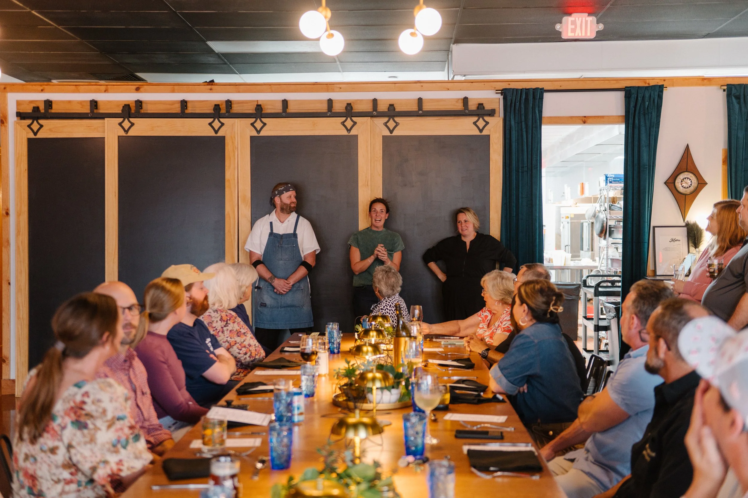People attending a presentation in a restaurant or event space, seated at a long dining table, with three presenters standing in front of a black chalkboard.