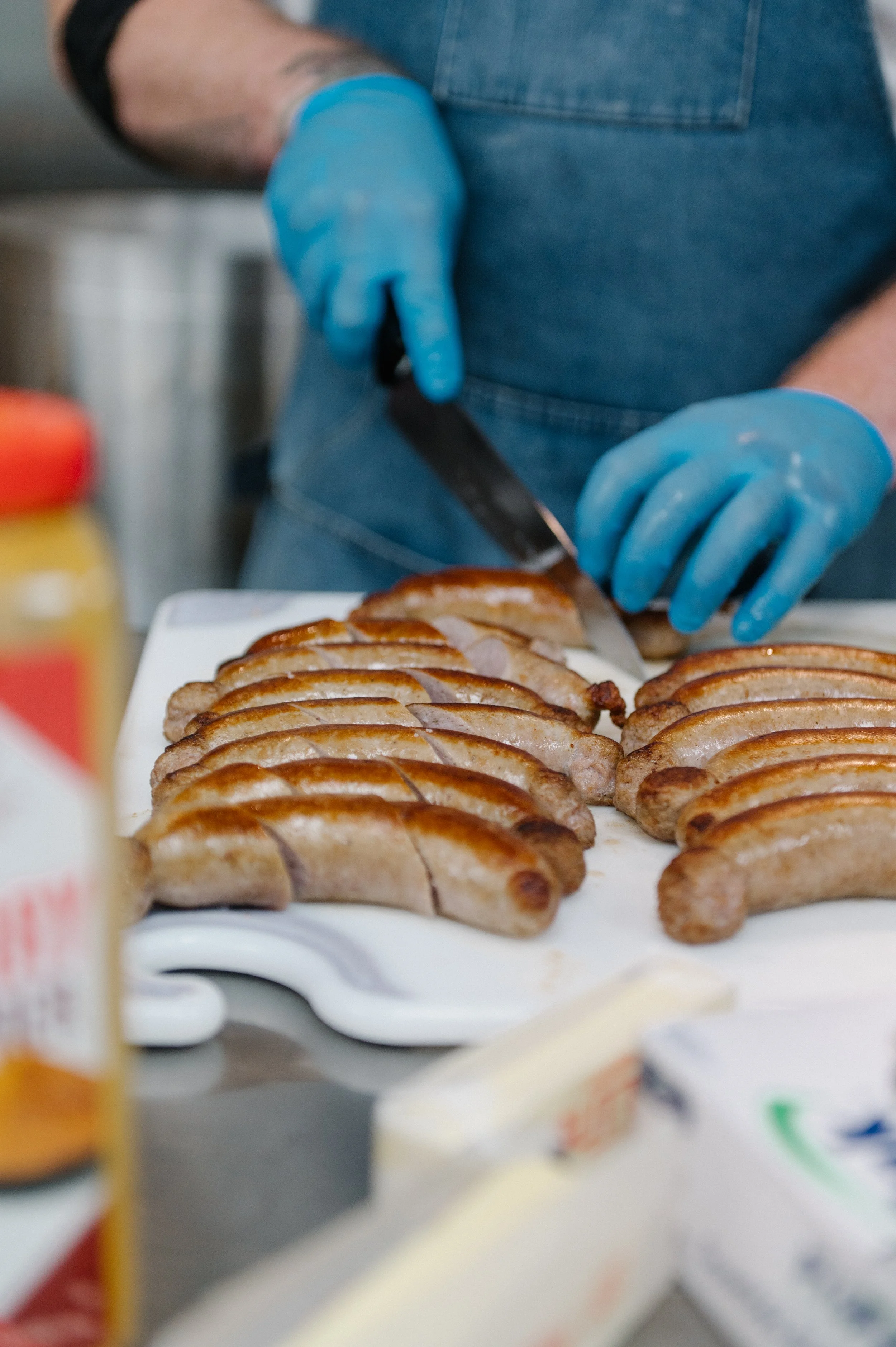 Person slicing grilled sausages on a white cutting board, wearing blue gloves and a denim apron, in a kitchen or food preparation setting.