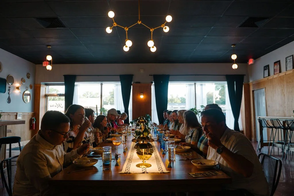 People dining at a long table in a restaurant with large windows, blue curtains, and modern lighting fixtures. Des Moines Wedding Rehearsal Dinner.