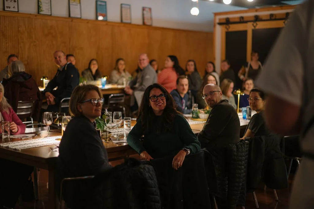 People sitting at tables in a dimly lit restaurant or cafe, with some looking towards the camera, and a person standing to the right in the foreground. Des Moines Dinner Party.