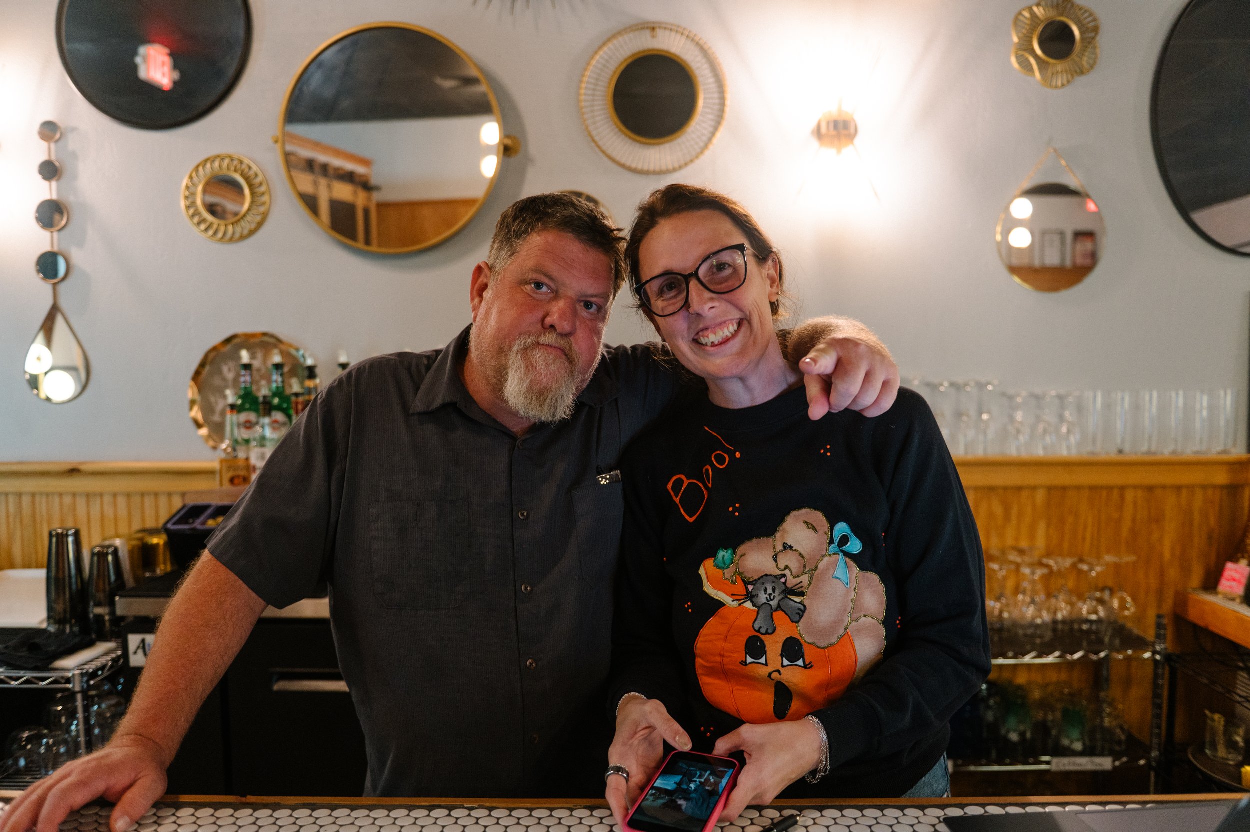 A man with a gray beard and a woman with glasses stand together behind a bar, smiling. The woman is holding a smartphone, and the man's arm is around her shoulder. They are in a restaurant or bar with decorative mirrors on the wall.