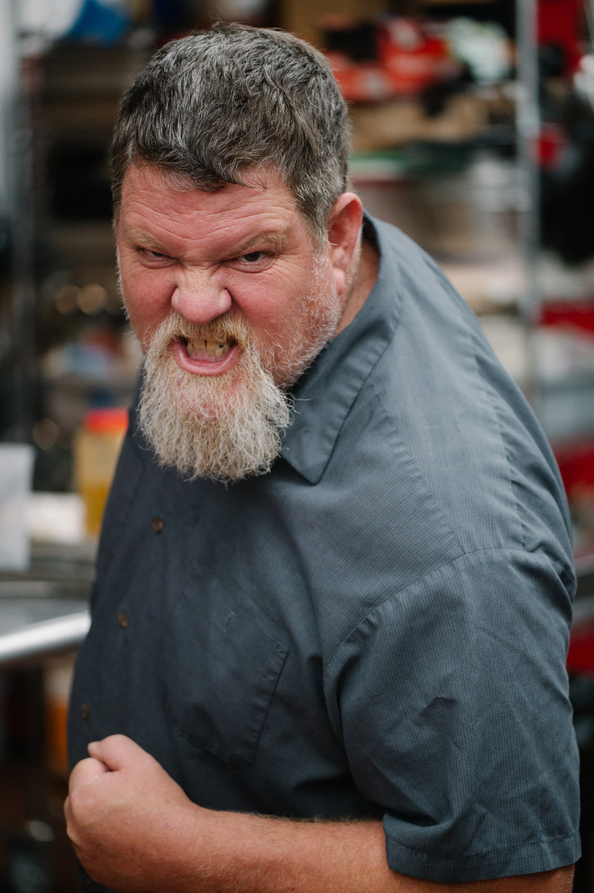 An angry man with a beard and short hair, wearing a gray button-up shirt, appears to be yelling or shouting in a kitchen or workshop setting.