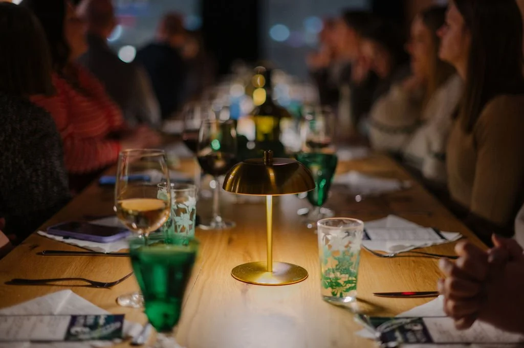 A dinner party with people sitting around a long wooden table, dimly lit, with glasses of wine and water, and a brass table lamp in the center. Des Moines event venue.