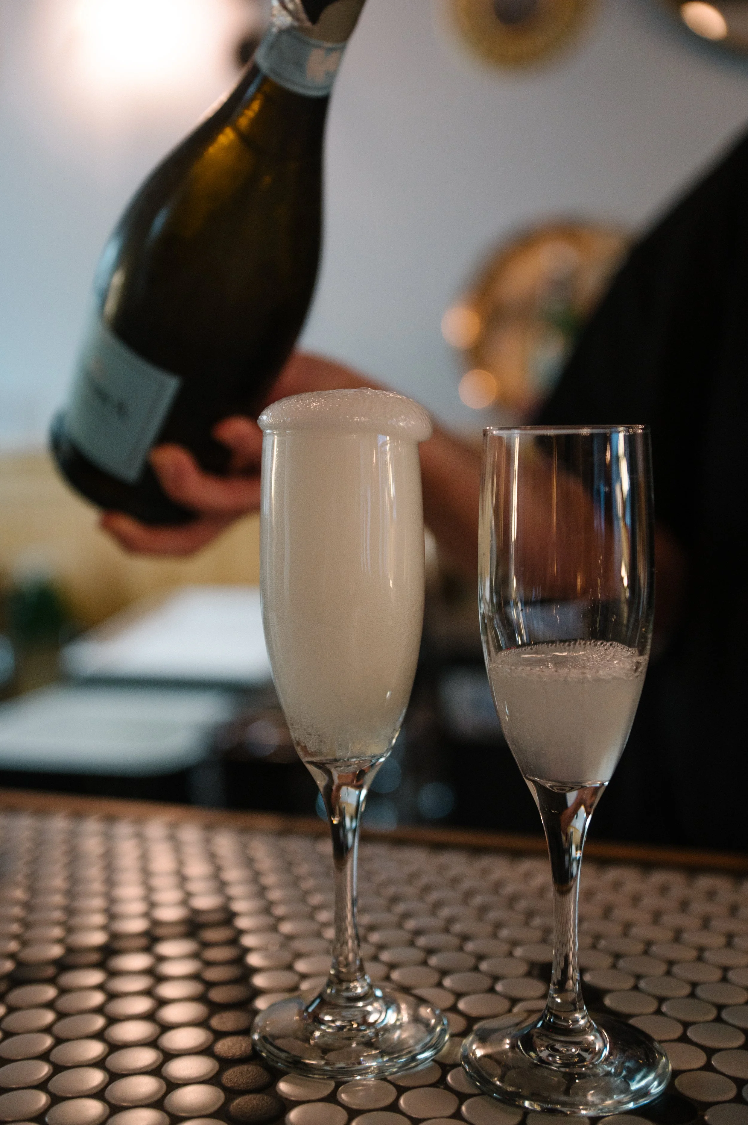 A person pouring champagne into a flute, with a second flute already filled and frosted, on a textured surface.