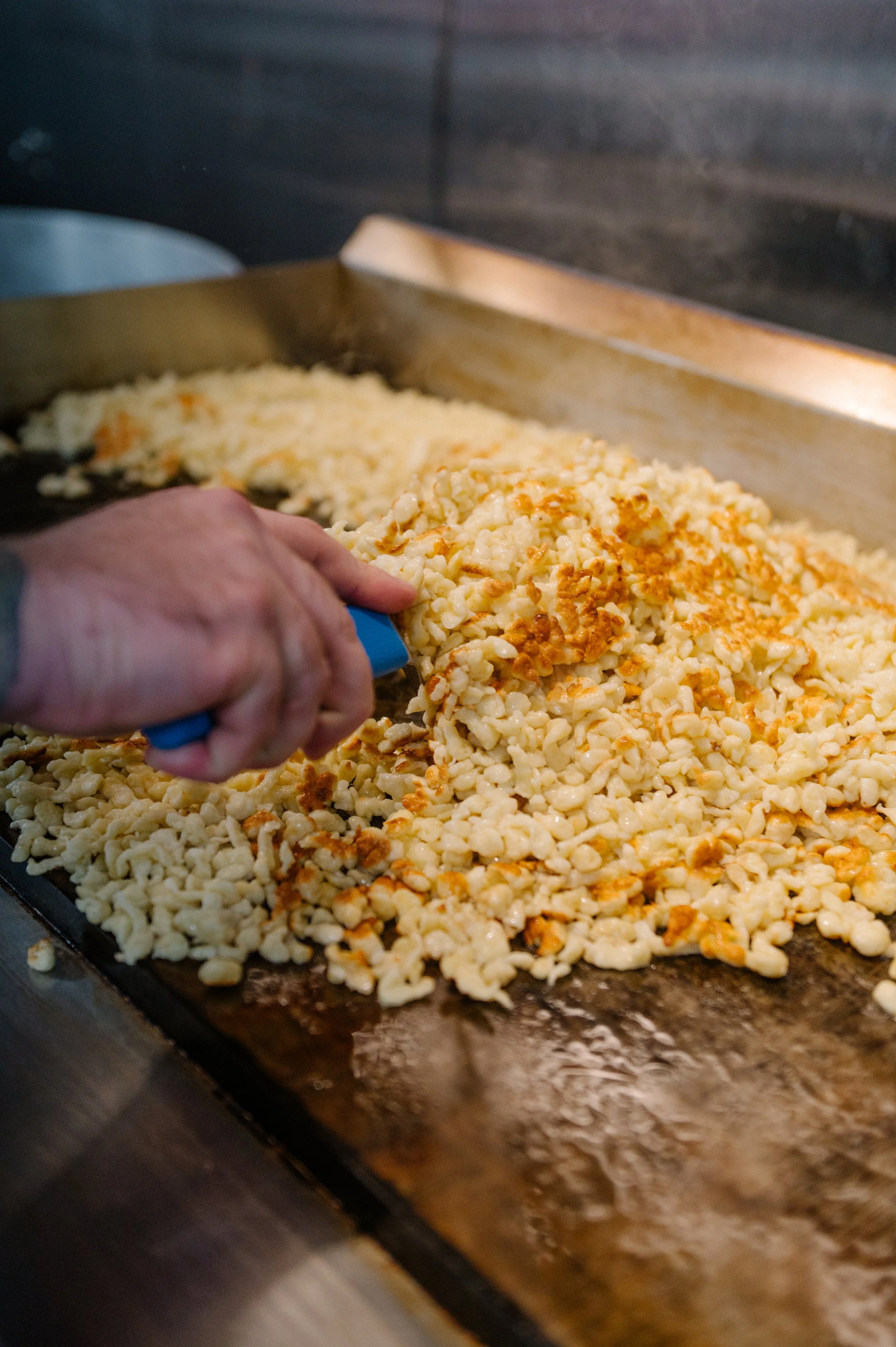 Food being cooked on a large flat griddle, with a hand using a spatula to stir or serve it.