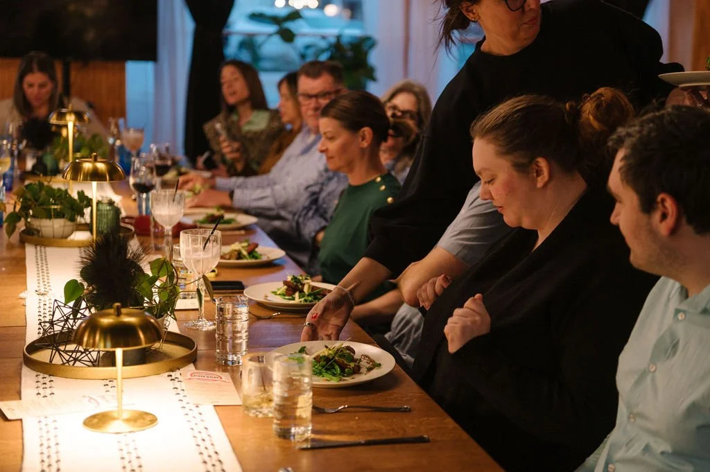 People seated at a long dinner table, with some plates of food and drinks, in a warmly lit room. Des Moines Engagement Party.