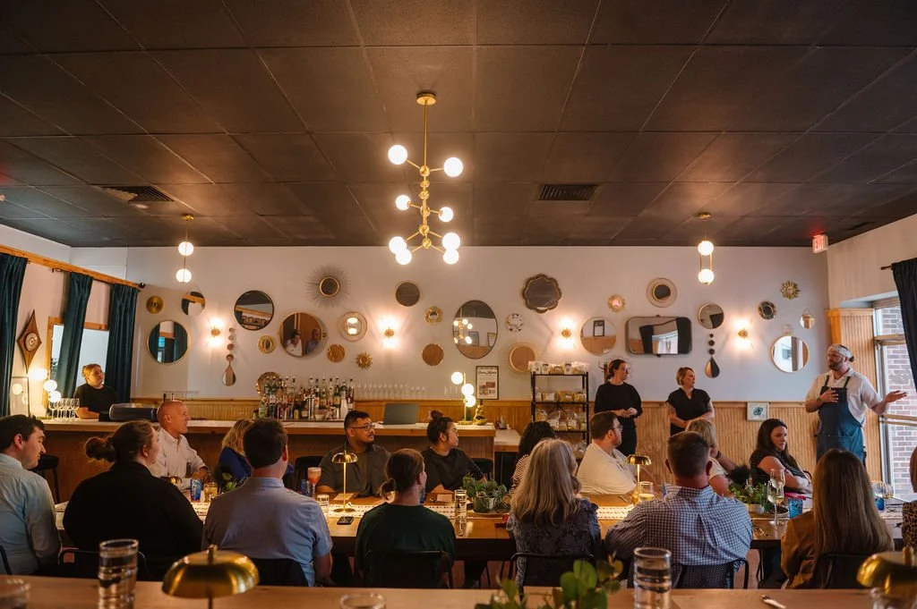 A group of people seated at a long table listening to a man speaking in a restaurant or bar with dark ceiling, modern lighting fixtures, and decorative mirrors on the wall. Des Moines Corporate Happy Hour.