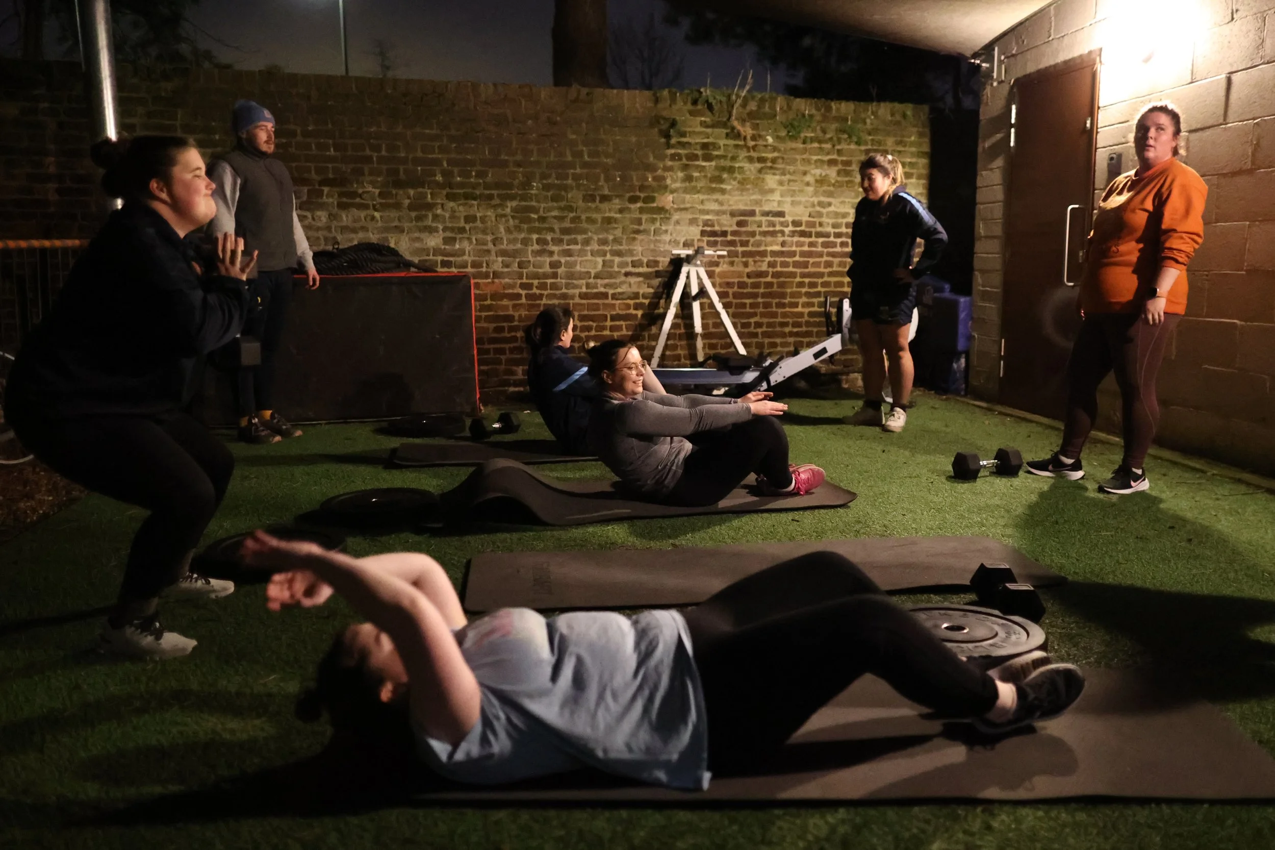 People exercising outdoors at night, with a woman lying on the ground doing leg lifts, others standing and observing, in a small yard with a brick wall and a wooden wall, fitness equipment scattered around.