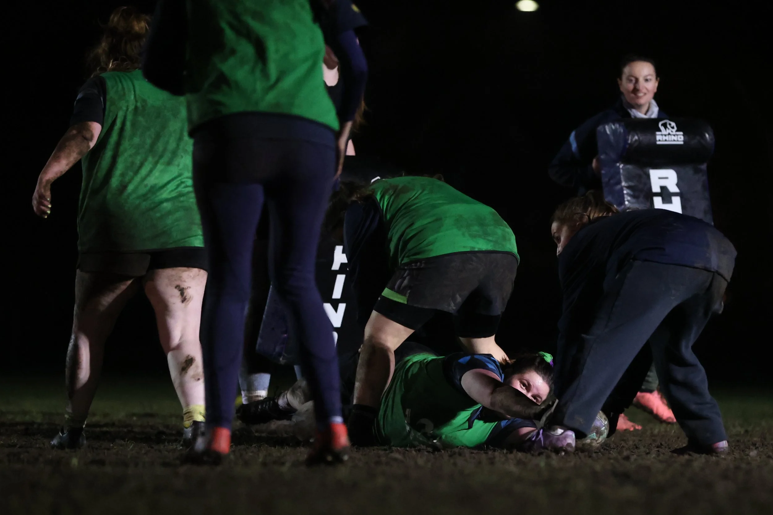 Women and men wearing sports clothing are participating in a rugby game on a dark field at night. Some players are bending down or standing, and one player on the ground is wearing a green jersey.