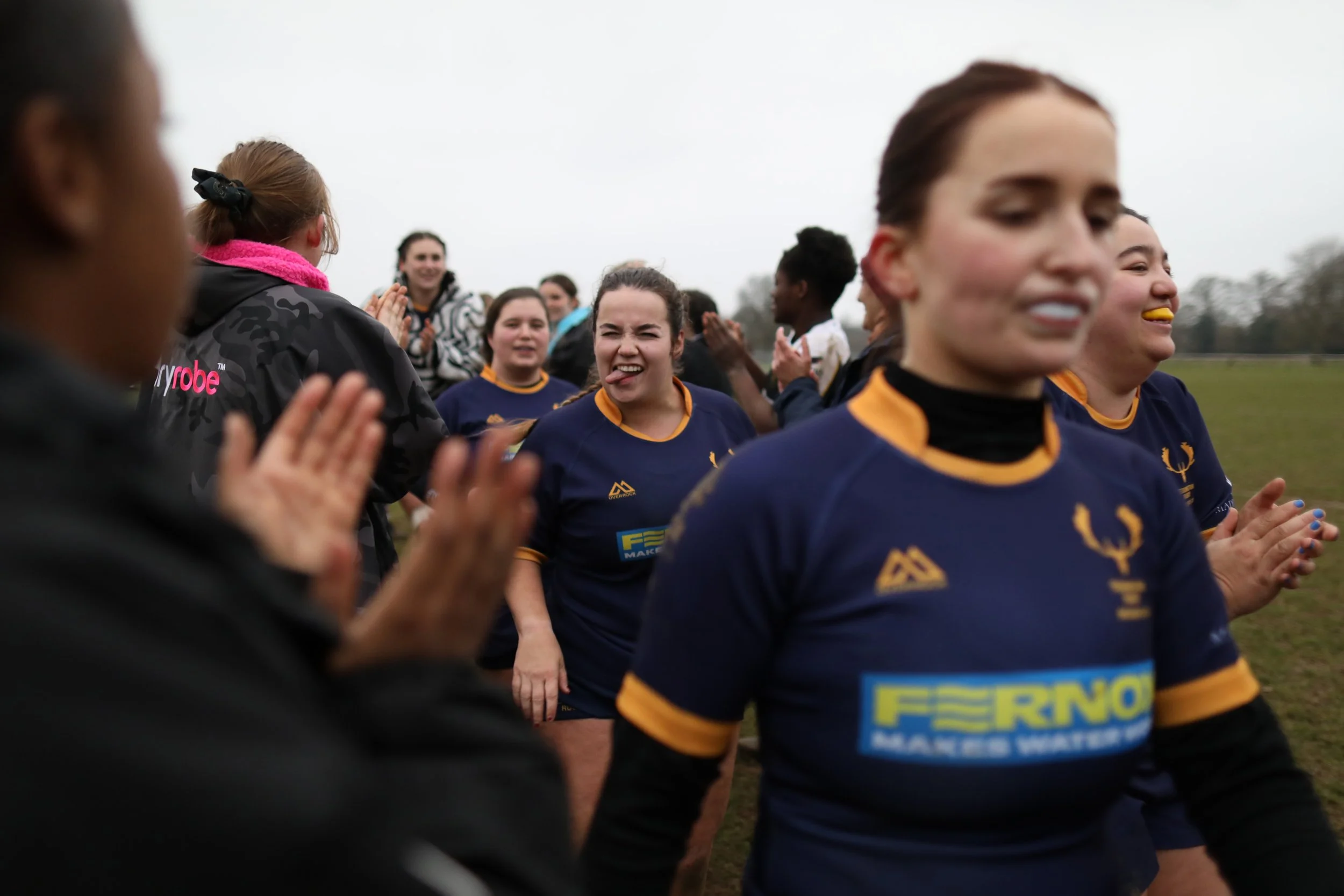 A group of female rugby players in blue and yellow jerseys celebrating and smiling on a grassy field, with some clapping and one player making a face sticking out her tongue.