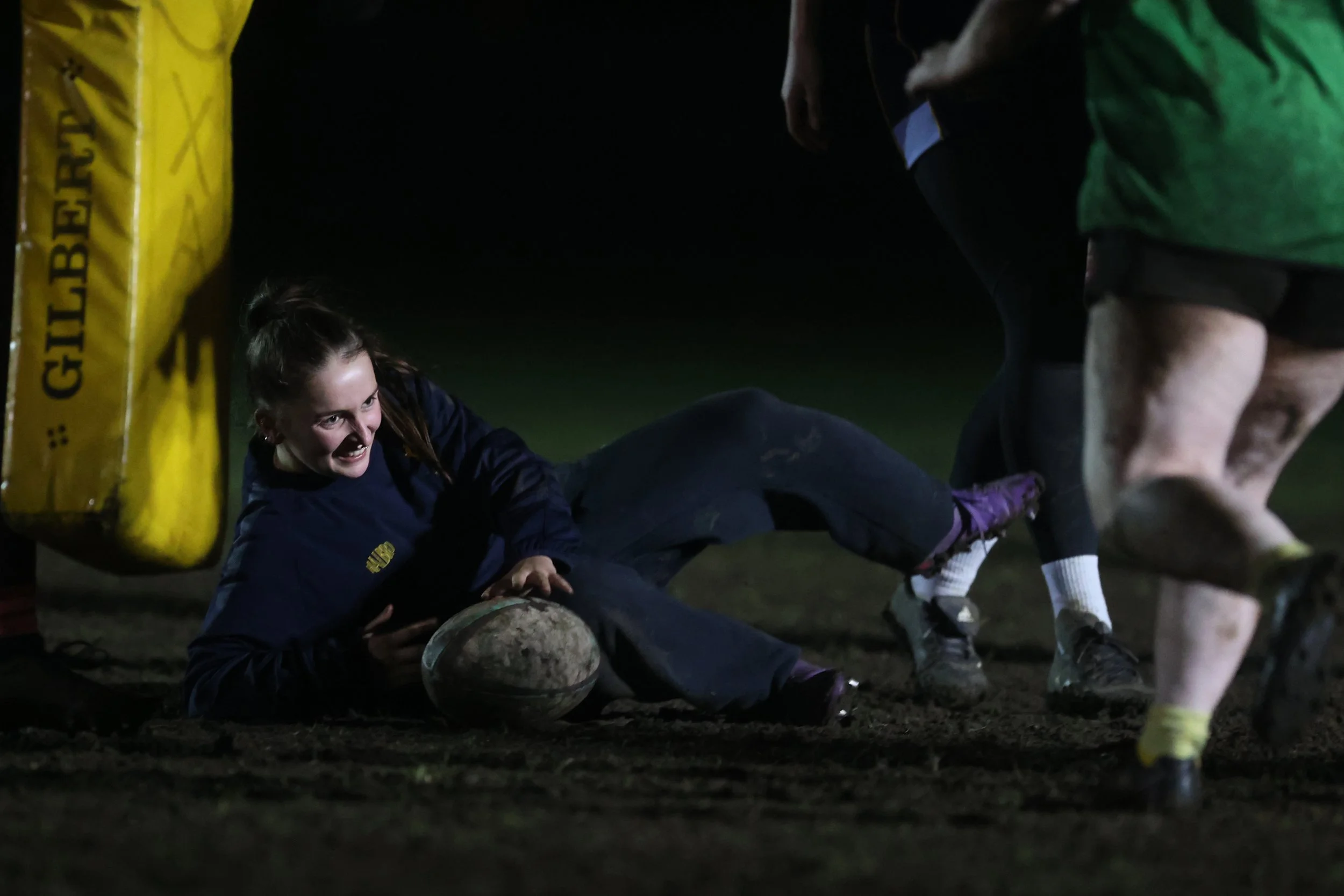 A person smiling while lying on the ground after falling during a nighttime rugby game.
