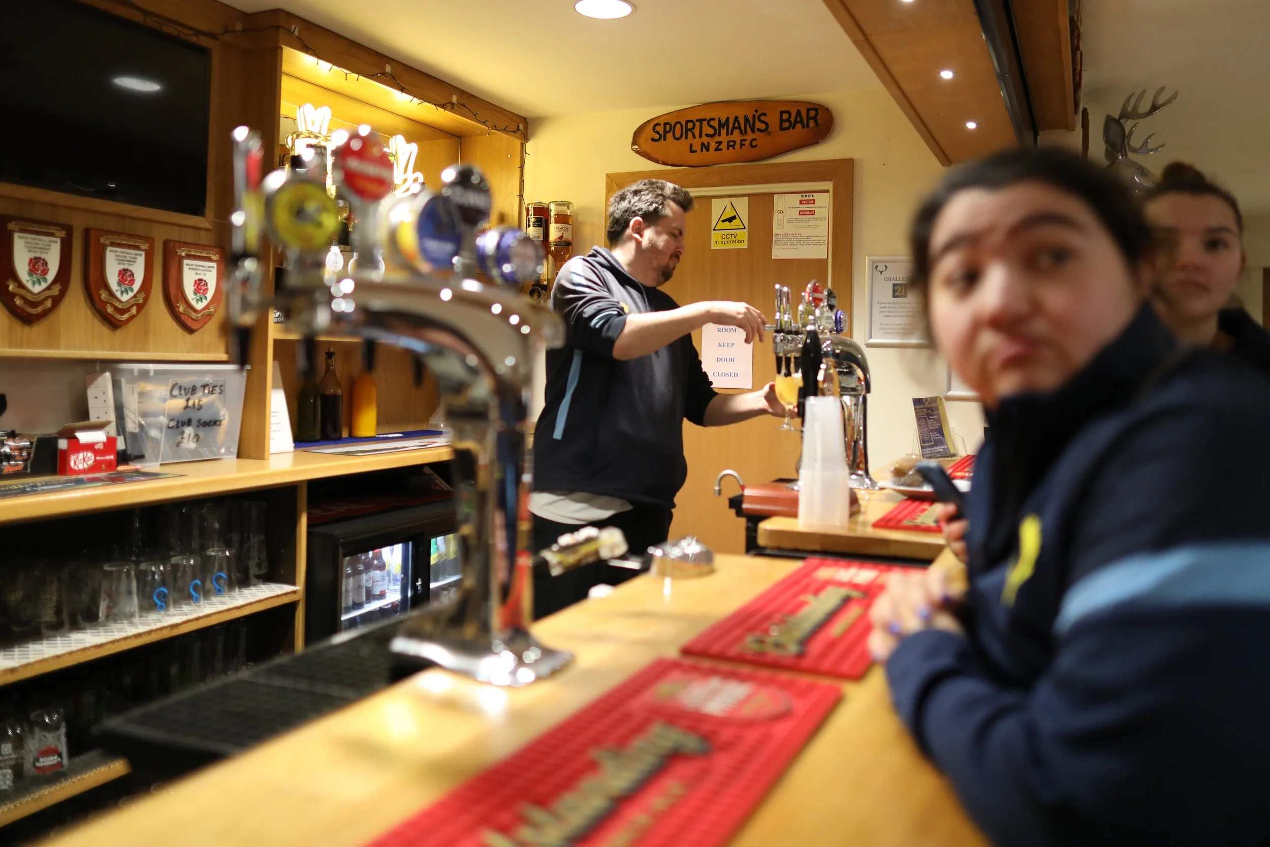 People at a bar counter inside a sports-themed pub. A bartender is pouring a drink, while two customers are seated in the foreground, one of whom is looking at the camera. The bar area has promotional signs, trophies, and wall decorations.