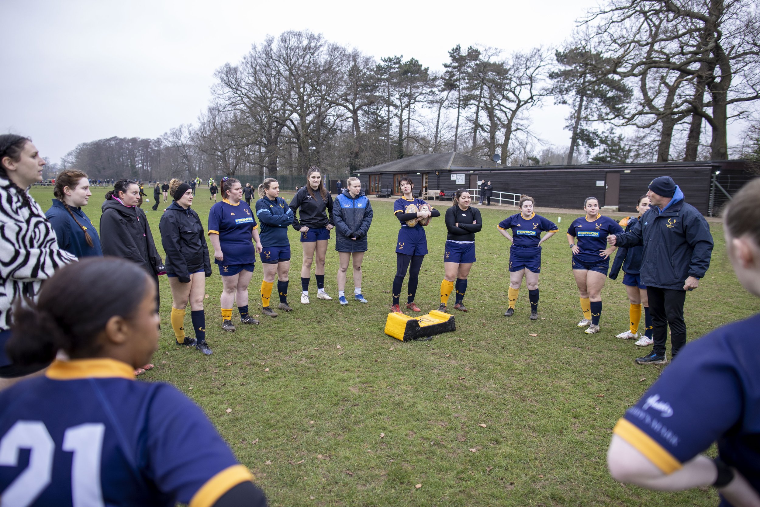 A group of female rugby players and a coach standing in a circle on a grassy field during daytime, with a wooden building and leafless trees in the background.