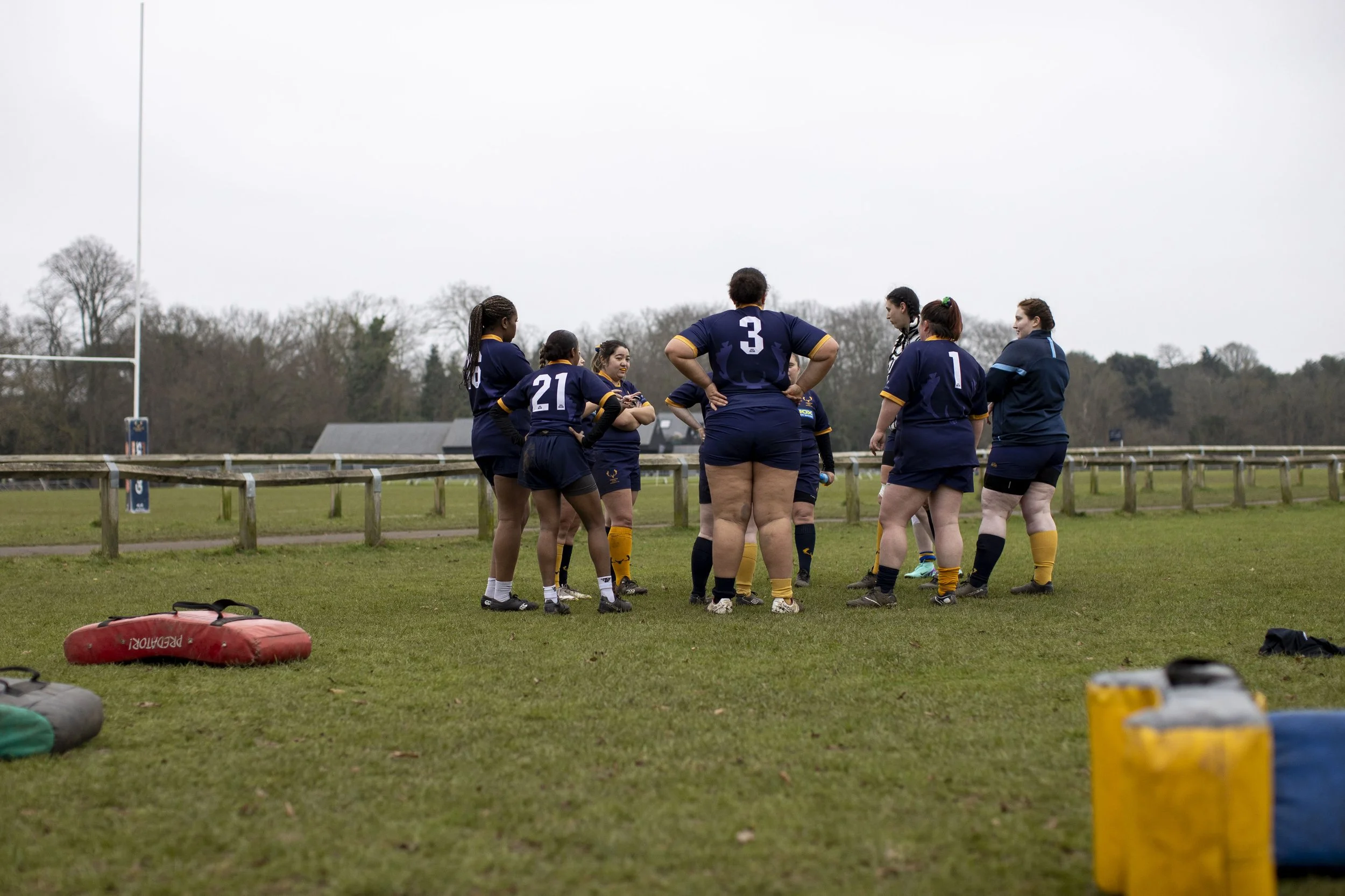 Women rugby players huddled together on the field during practice or a game, with rugby gear and equipment visible on the grass, and a rugby goal post in the background.